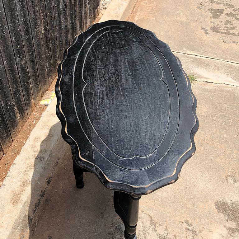 Black oval scalloped edge side table. Featuring an oval wood top with scalloped edges. Small detail carving around the perimeter. Some wear to the top as shown in photos. The base features 4 legs with spindle design. All four attached by an