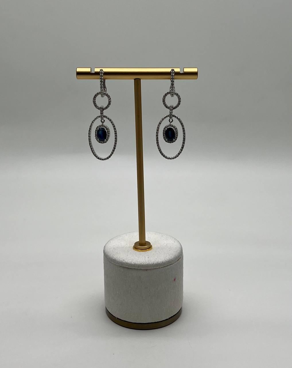 Elegant and contemporary, these exquisite drop earrings feature a captivating design of interlocking circles adorned with shimmering diamonds. At the center, a deep blue sapphire is encircled by a halo of sparkling accent diamonds, creating a
