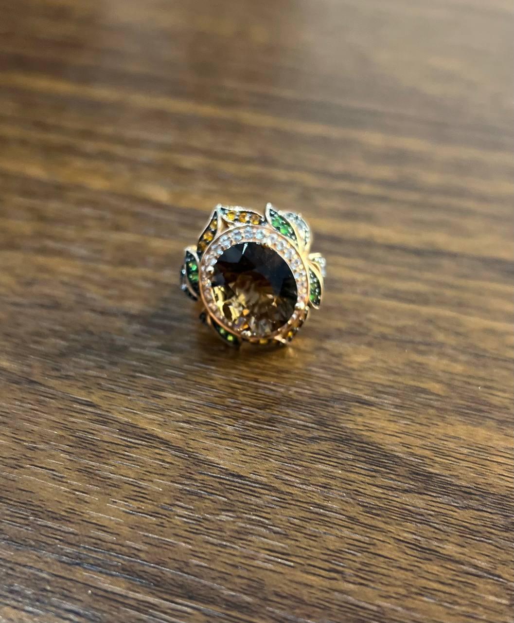 Exquisito Anillo Multipiedra con Cuarzo Ahumado y Detalles de Gemas. Este impresionante anillo presenta un gran cuarzo ahumado facetado en su centro, rodeado de un halo de brillantes diamantes blancos y gemas de colores. El intrincado diseño incluye