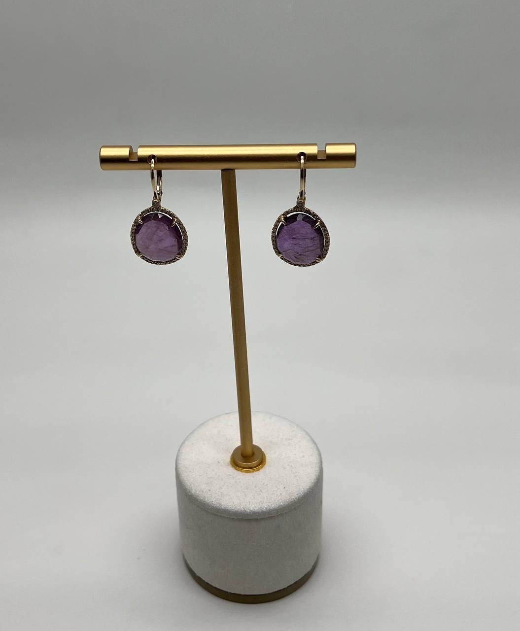 Elegantes pendientes en forma de gota con gemas de amatista púrpura de forma ovalada. Las gemas tienen un matiz rico y translúcido con inclusiones naturales que añaden carácter. Engarzada en un marco de oro pulido rodeado de diamantes color champán,