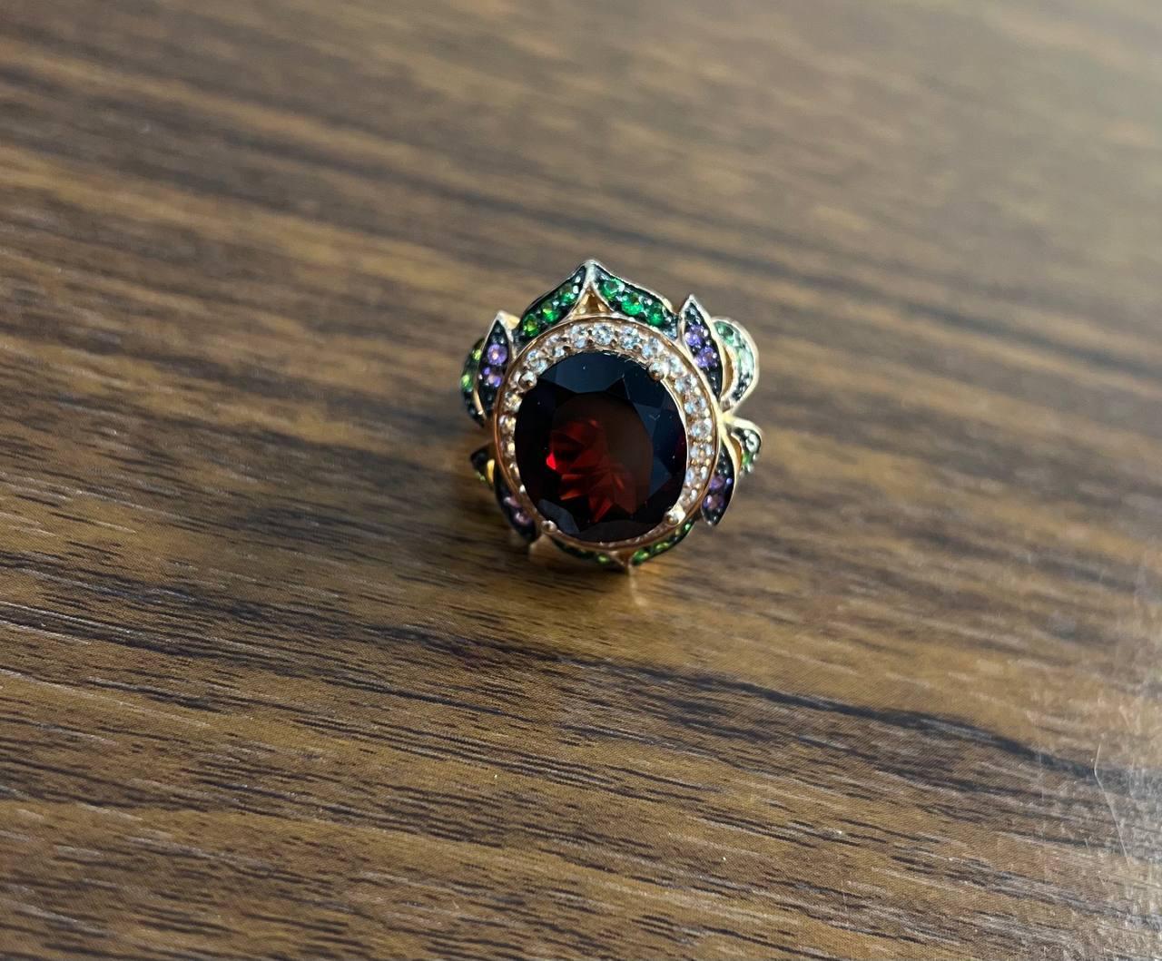 Un llamativo anillo de varias piedras. Esta exquisita pieza presenta una rica banda de oro rosa adornada con un impresionante conjunto de piedras preciosas. La pieza central es un granate ovalado de color rojo intenso, sujeto con seguridad por un