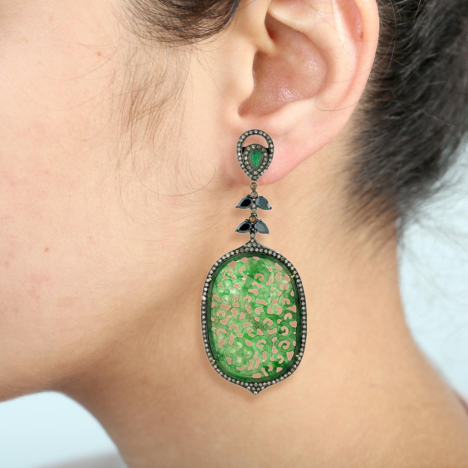 Gorgeous oval-shaped carved jade dangle earrings with emerald and spinel accents and shimmering diamonds. 

18k:1.71g,
Diamond:3.5ct,
Silver:20.65gm,
Emerald:0.7ct,
Jade-41.85,
Spinel-4.10
Size: 90X36MM