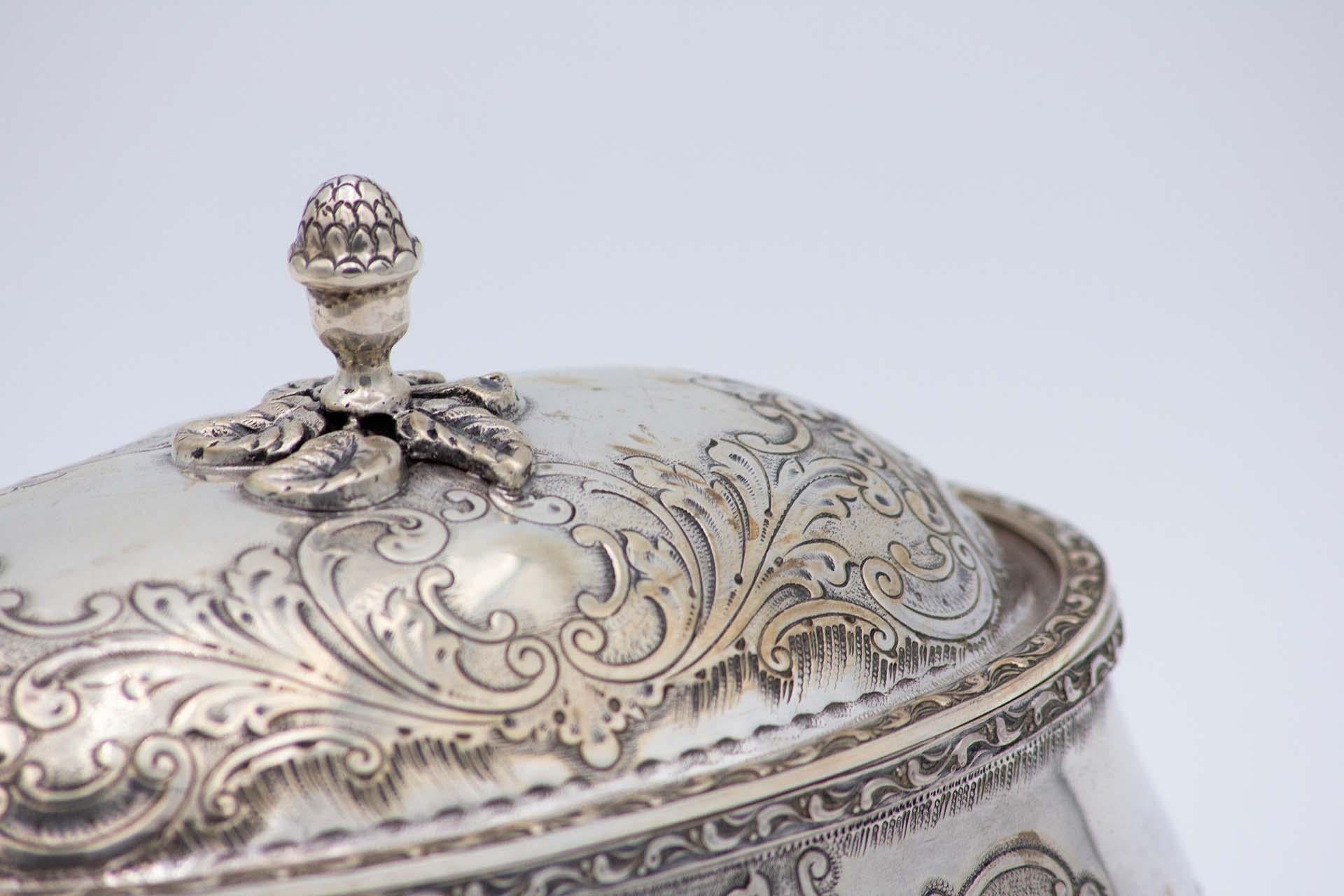 Description: Oval-shaped silver scullery with embossed floral motifs, removable lid knob and resting on four acanthus leaf-shaped feet. Metal: Silver. Origin: Portuguese. Brand: Coelho. Weight: 624.01 grams. Dimensions: 18.5cmx14cm. Content: