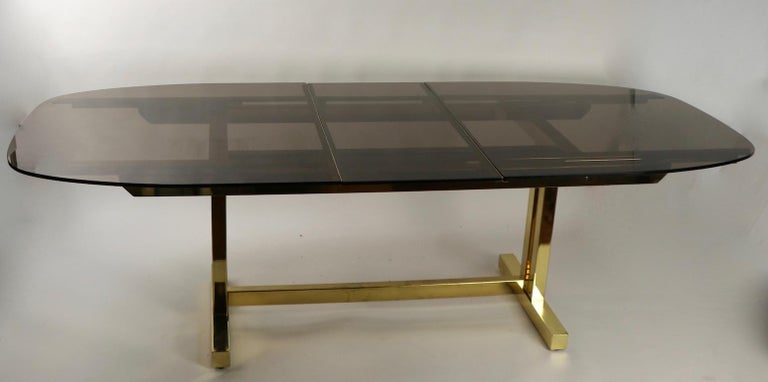 Impressive Postmodern, Art Deco Revival dining table of brass and smoked glass. This unusual table is extendable, having a 18 inch W leaf which stores under the glass surface when table is closed. Total W with leaf in place 95.25 x 77.5 W when