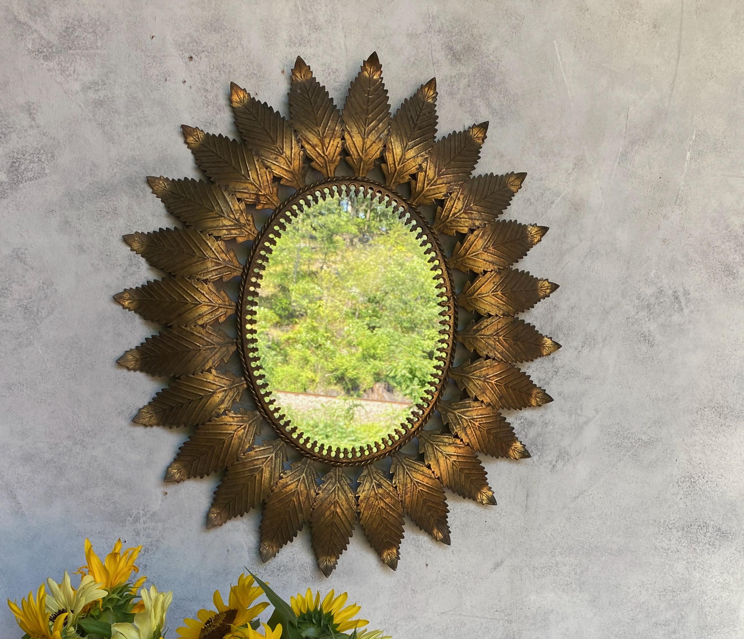 An oval gilt metal sunburst mirror with classical stylized leaves radiating out  from an interior border, representing a modern trefoil design. We recently added a felt backing to the mirror to give it more protection and a finished look. Spanish