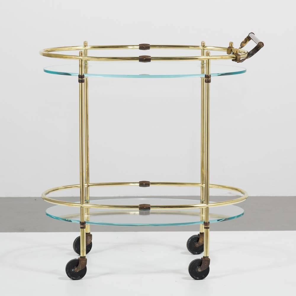 Oval polished and patinated brass framed bar cart on two tiers with glass shelves set on castors, 1960s.


