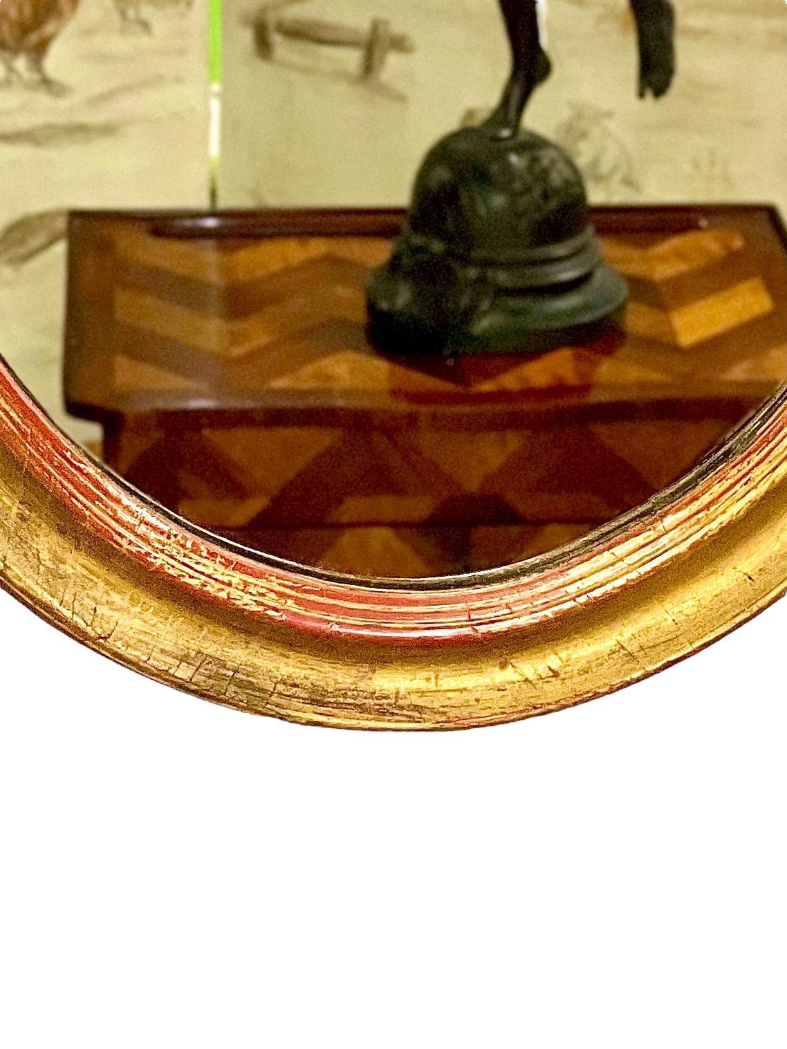 Ce magnifique miroir mural ovale français présente un cadre en bois doré sculpté d'une simplicité classique, avec un contour de moule en escalier et concave entourant la plaque du miroir. Avec une patine joliment vieillie, les nuances rouges du bois