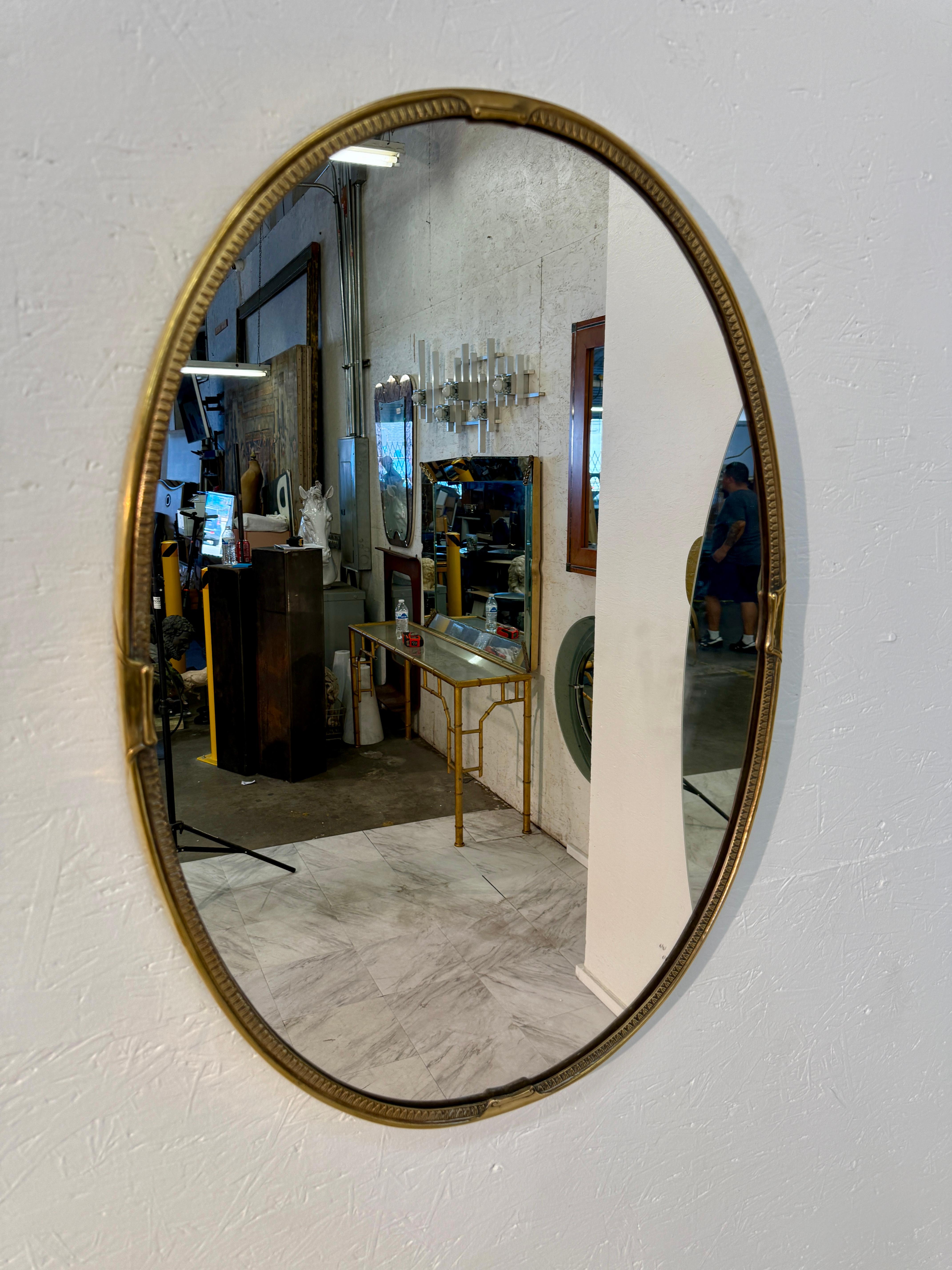Espejo de pared ovalado vintage encerrado en un marco de latón macizo. El marco presenta intrincados detalles grabados a lo largo del borde interior, con detalles decorativos en forma de cinta situados en la parte superior, inferior y lateral, que