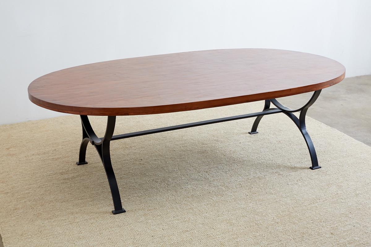 Grand oval wooden dining table featuring a curule leg style iron base. Hand-crafted of thick wooden planks with a hand-hewn finish on top and wood peg construction. Supported by a thick iron bar base with a pair of curule form legs conjoined by a