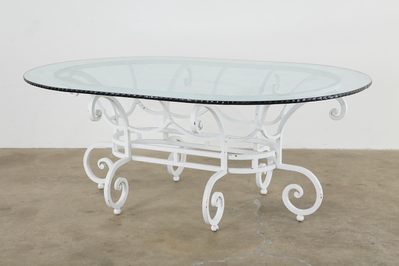 Impressive oval garden dining table featuring a large wrought iron base. Crafted from thick rods in a basket form with scrolled ends. The base is supported by six scrolled legs ending with ball feet. The large base is topped with a thick pane of