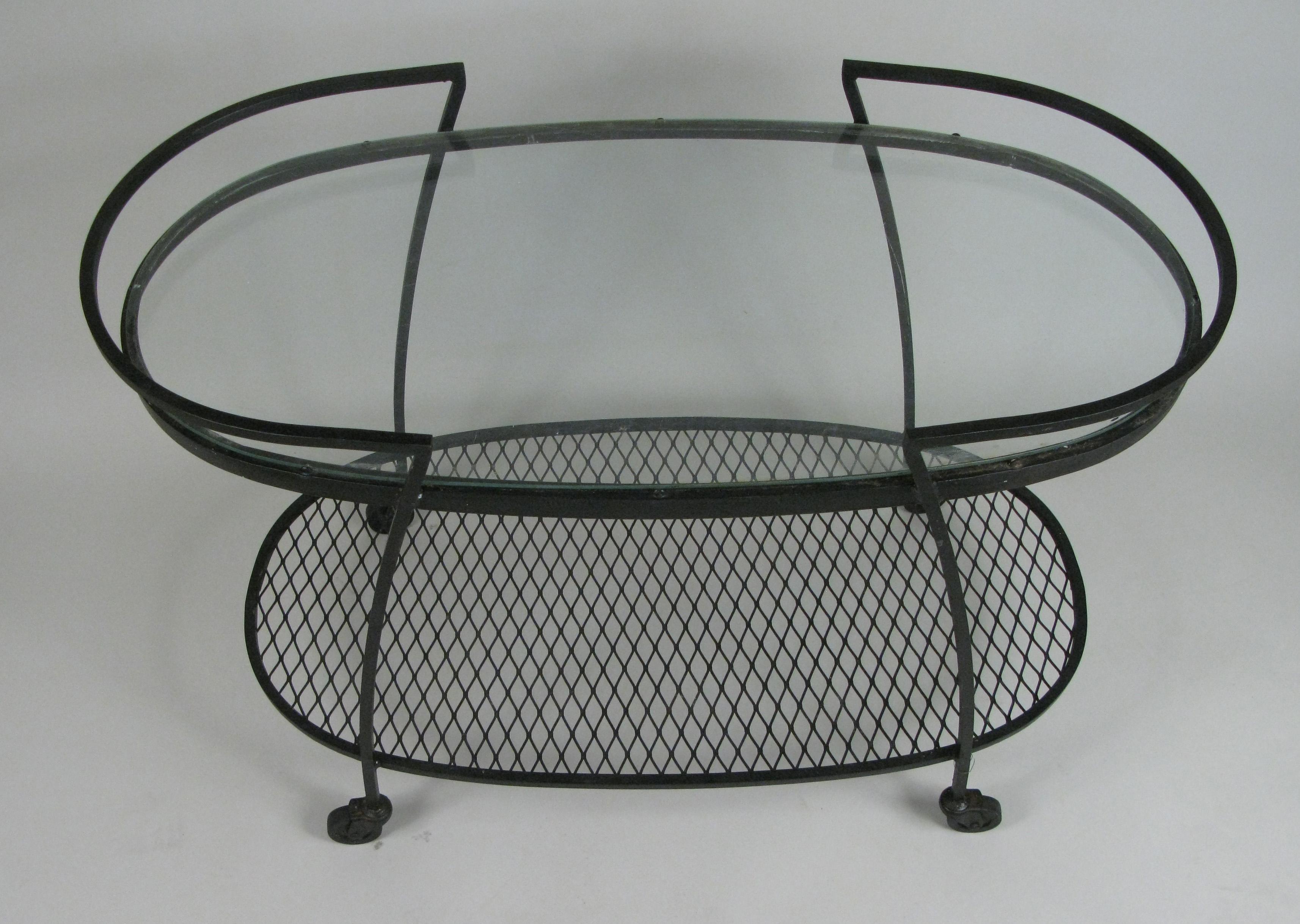One of the best of Woodard's series of wrought iron rolling bar carts from the 1950s, this oval design from the Pinecrest collection has a low steel mesh shelf, and a large glass oval top. The handles curve around the ends and provide a rim to keep
