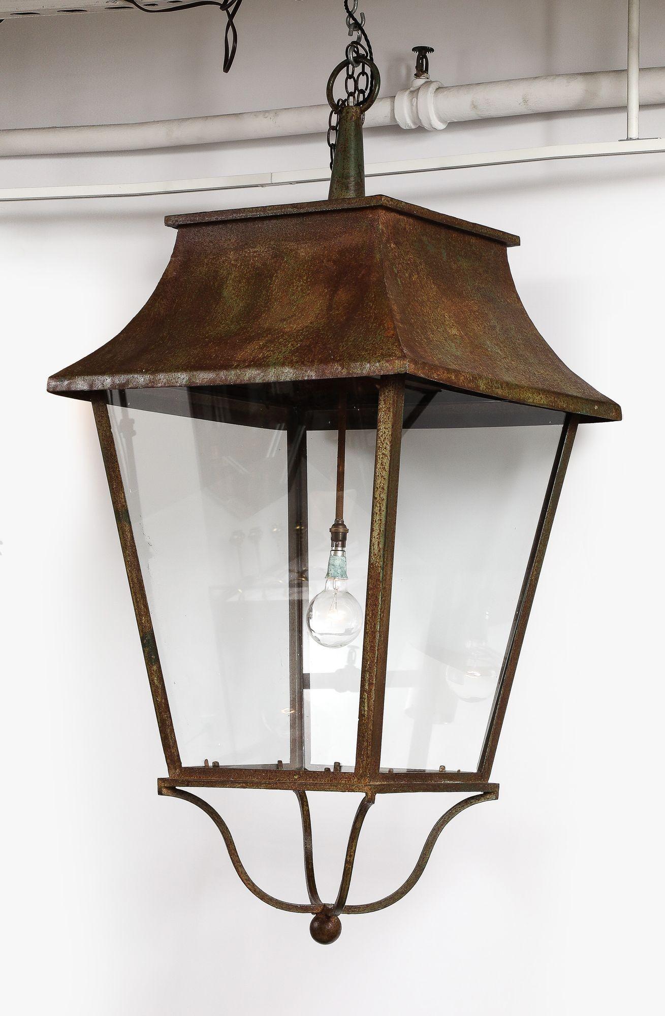 A magnificently scaled English country house hall or stable lantern in patinated iron with integral hanging loop above curving hood and glazed, canted sides, with shaped iron base ending in a center ball. Electrified with central hanging fixture.