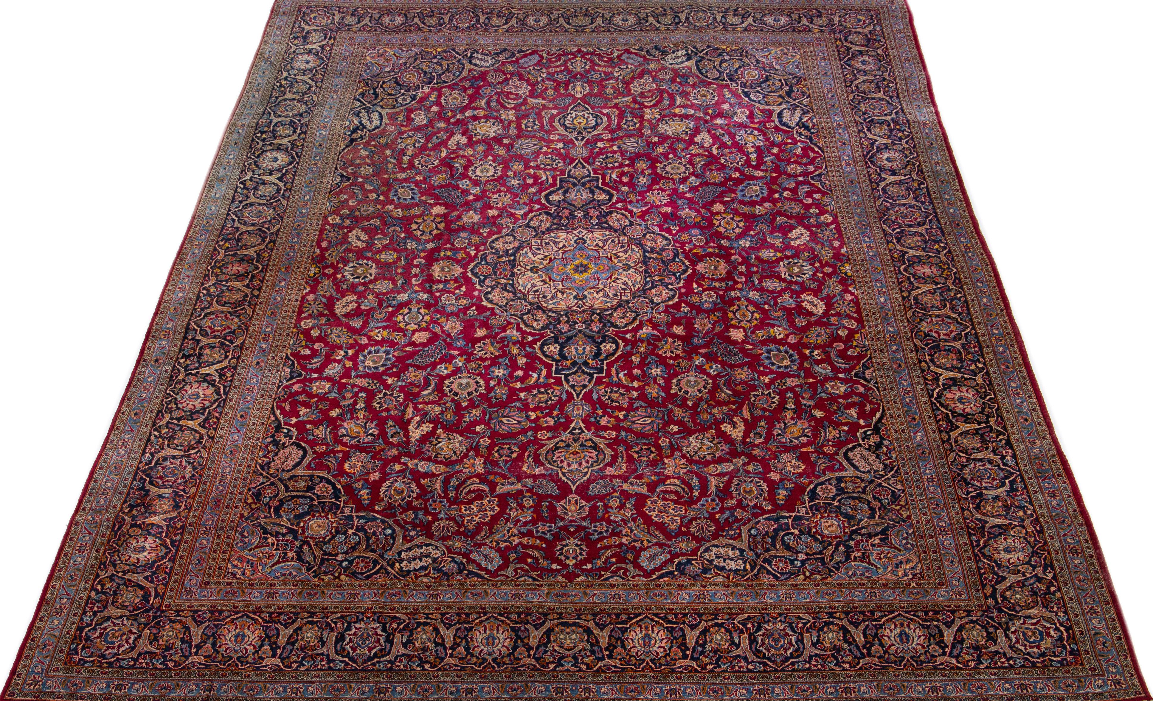 This antique Kashan wool rug boasts an exquisite design, featuring a red color field and a distinguished blue frame adorned with stunning multicolored floral accents. Its oversized dimensions add to the rug's allure, making it a true masterpiece of