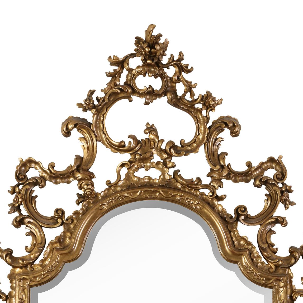 A large scale French rococo style mirror with ornate gilt detail. Carved scroll, leaf and floral design decorate the mirror border. The mirror itself has an arched shape at top and square corners at the base. The open work, carved and gilt frame has
