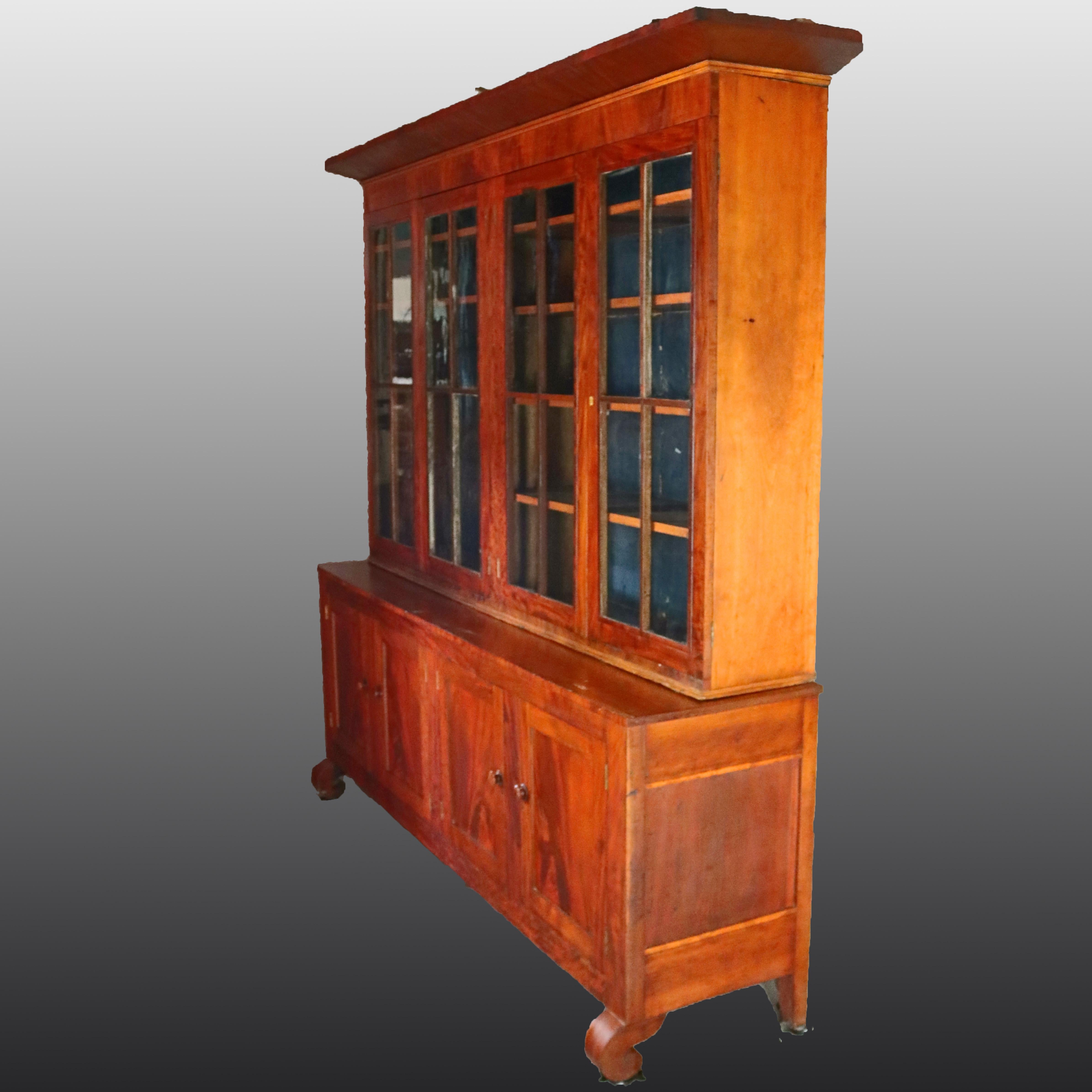 An oversized antique American Empire breakfront cabinet offers deeply striated flame mahogany with upper haing four glass doors opening to shelved interior with lower cabinet having four doors also opening to shelved interior, raised on scroll form