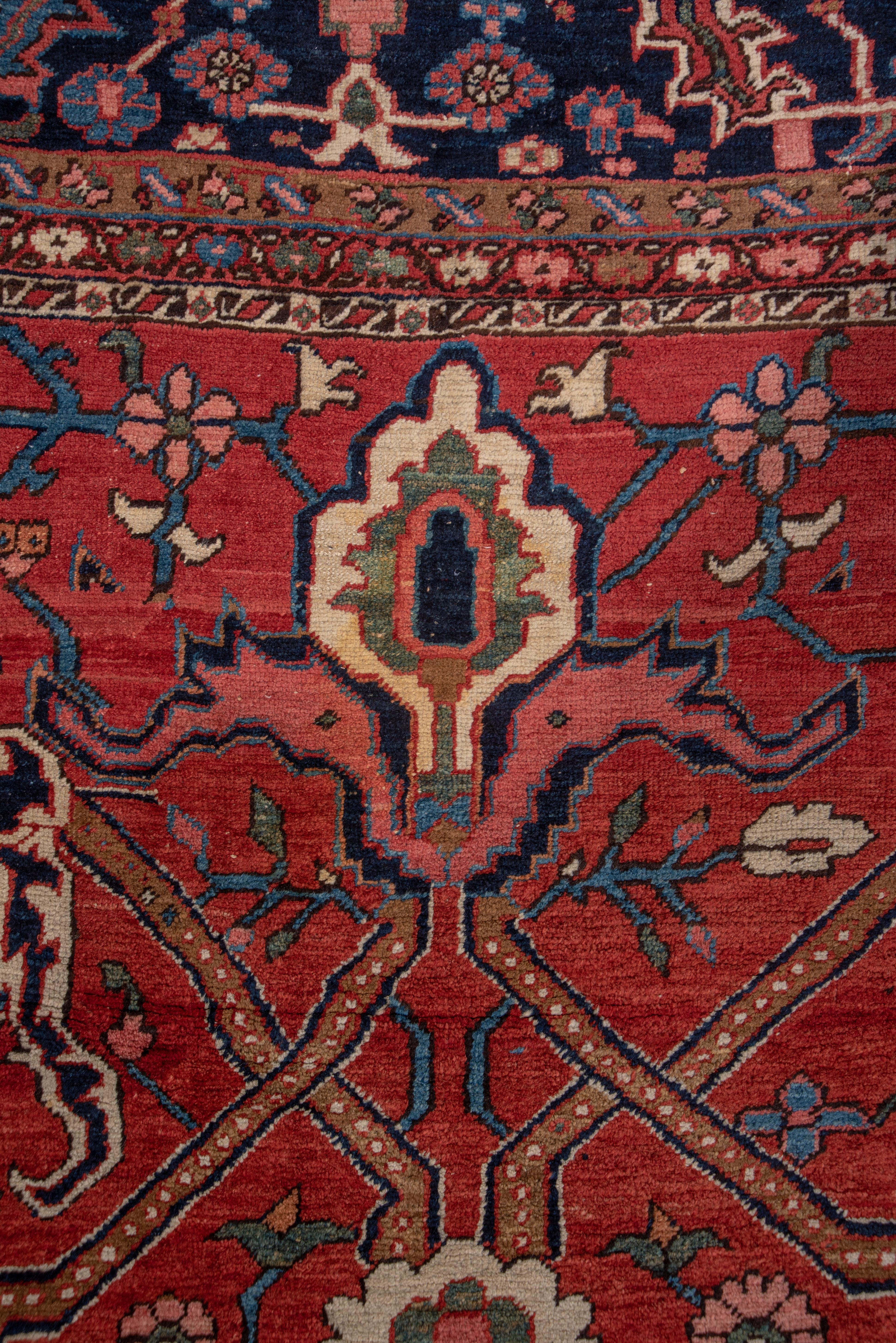 This magnificent NW Persian rustic oversize carpet from circa 1910 displays a warm madder tomato red field with a strong dark blue strap work pattern organizing winged escutcheons and palmette pendants around a central multi-petal rosette. The scale