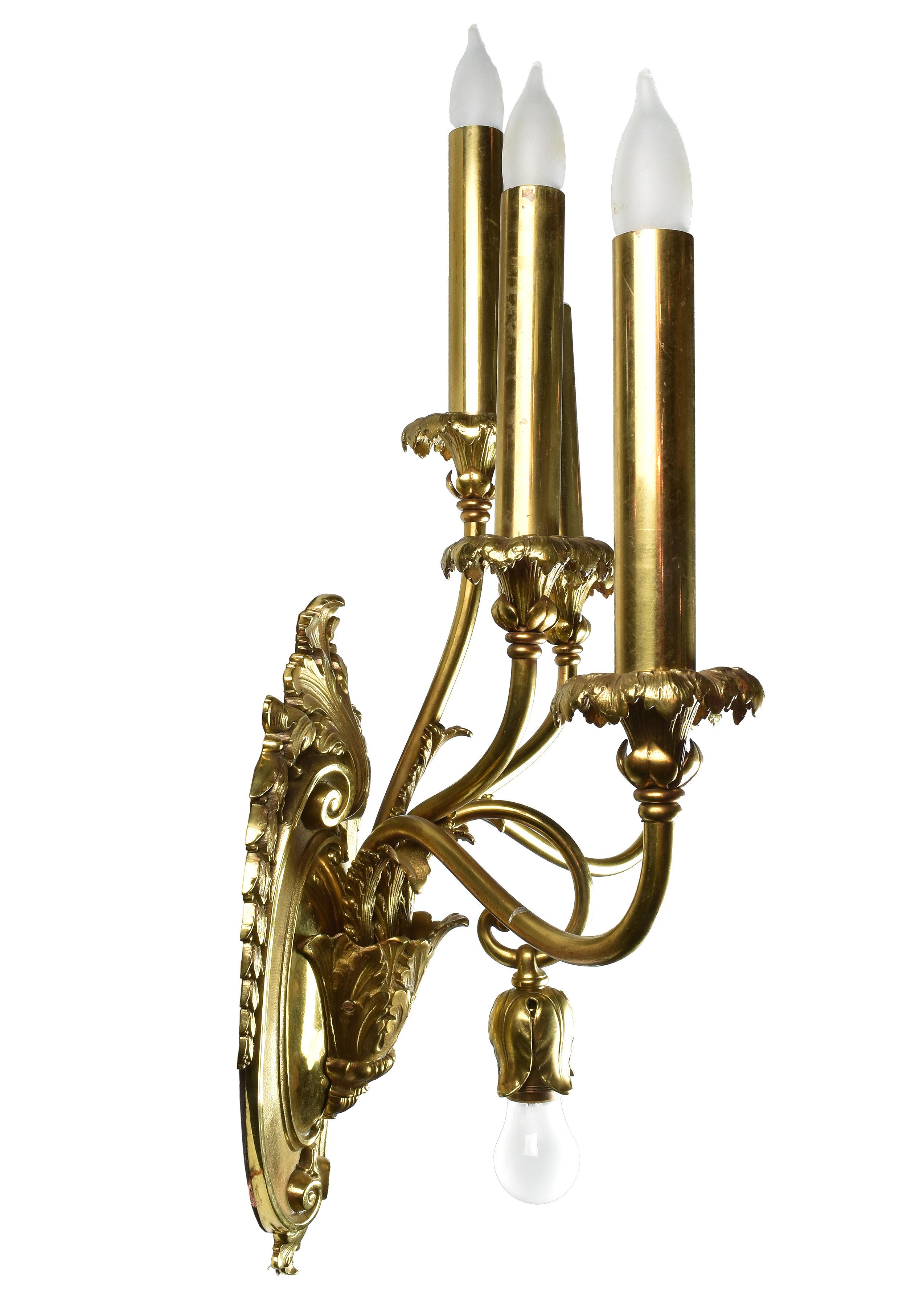 This finely crafted pair of oversized brass sconces features intricately detailed floral accents within the brass, 5 looping candle arms and an additional upside-down hanging bulb within the center of the fixture.