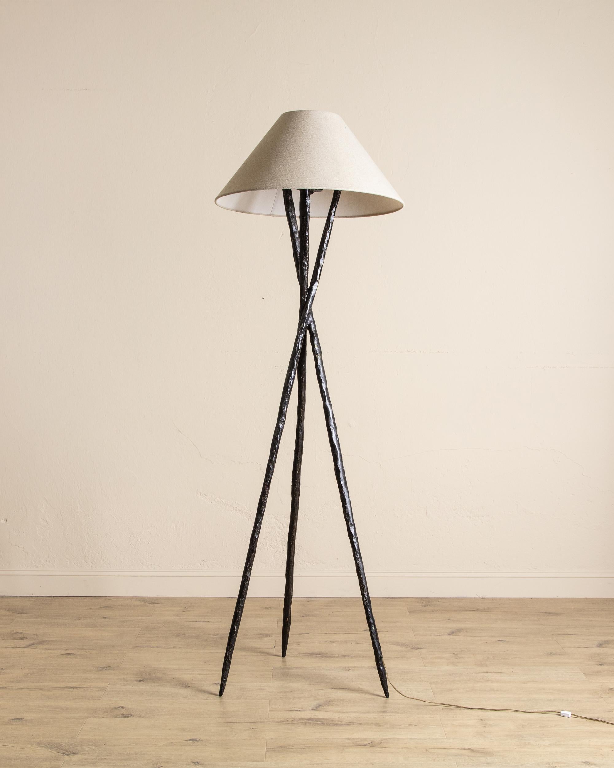 This striking floor lamp features a sculptural tripod base crafted from hand-forged metal, offering a refined blend of modern design and artisanal character. The hammered texture of the legs adds depth and visual interest, giving the piece a