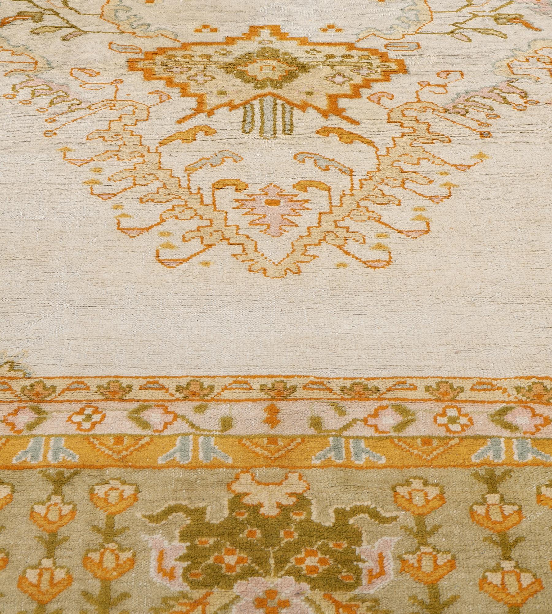 This antique Oushak rug features an ivory field with a central lozenge medallion containing a central dusty-pink and sandy-yellow floral lozenge issuing a variety of floral stems surrounded by a band of delicate floral vine with a bold burnt-orange