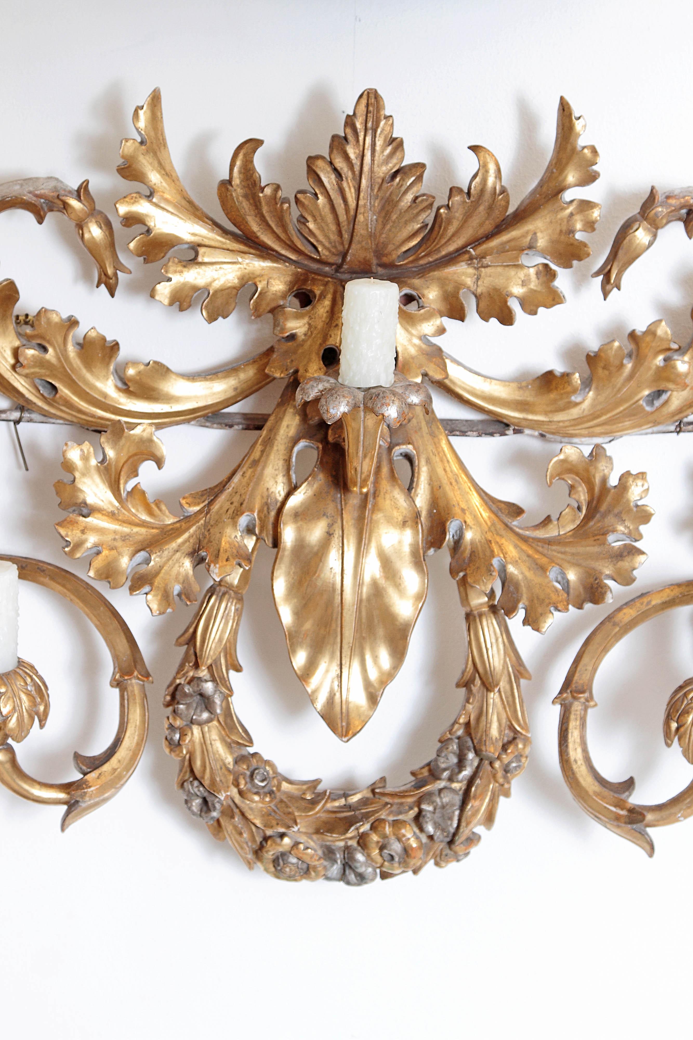 An oversized / large seven-arm gilt carved-wood and gesso wall sconce, gilded gold with silver accents, Baroque-style, undulating acanthus leaves swirls and curve to make the various candle arms, a floral wreath at centre bottom, large single