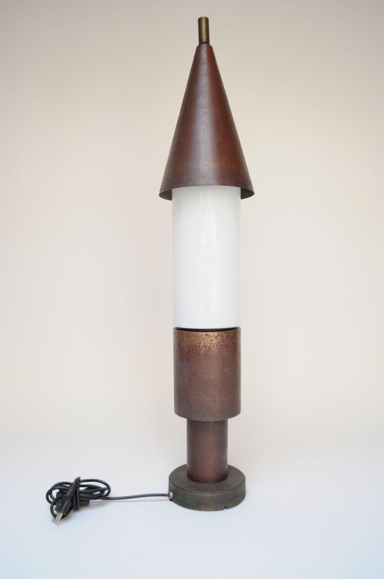 Lámpara de sobremesa alta, visualmente llamativa, con pantalla triangular patinada de cobre y bronce con remate de latón y vástago inferior de cobre que flanquea una columna central de cristal opalino, todo ello apoyado en una base redonda de acero