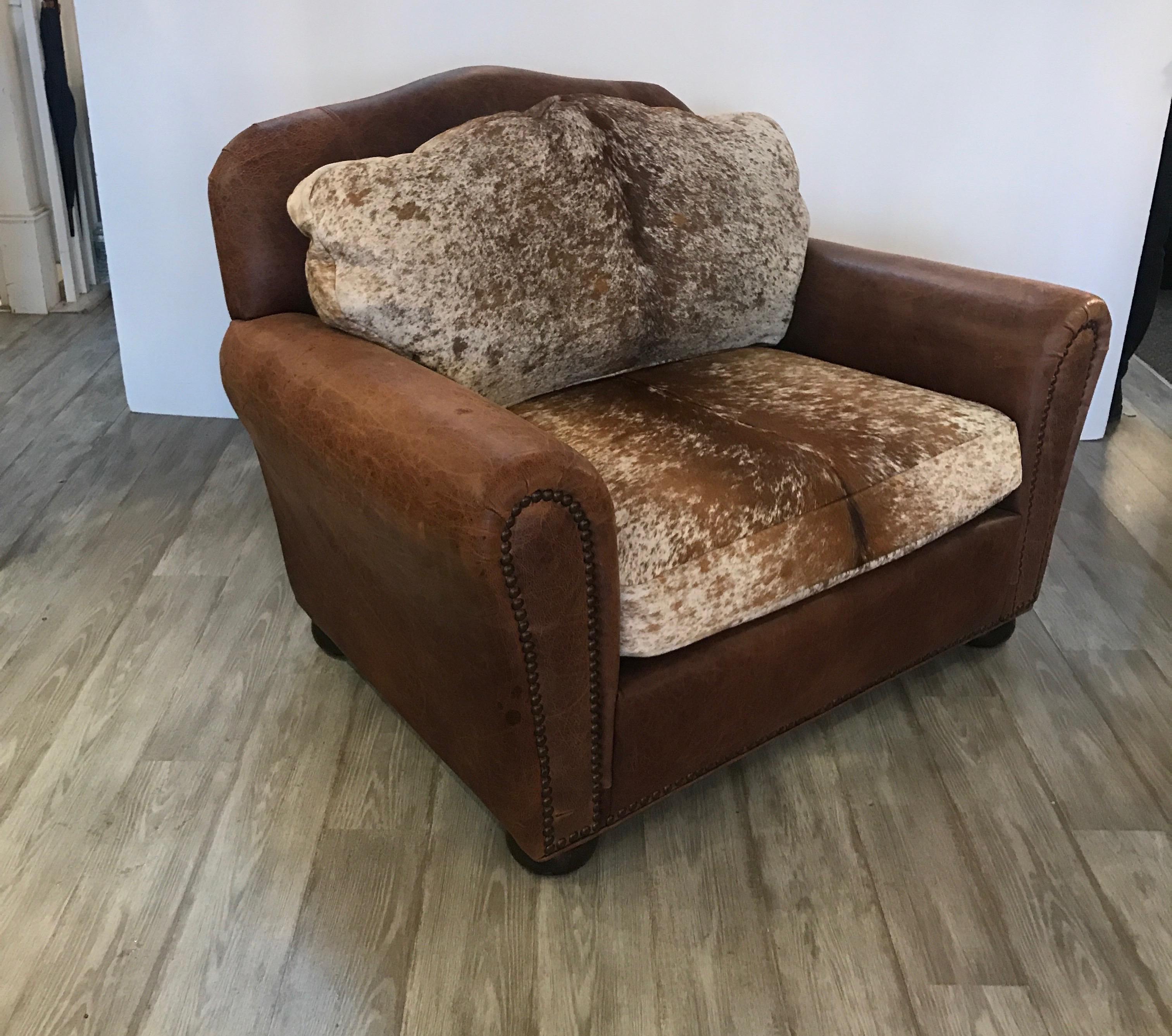 A large leather and cow hide club chair. The medium brown leather with intentional light distressing with back and seat cushion with cow hide.
The front with antiqued brass nailhead trim. Extremely roomy and comfortable in the style of Ralph Lauren.