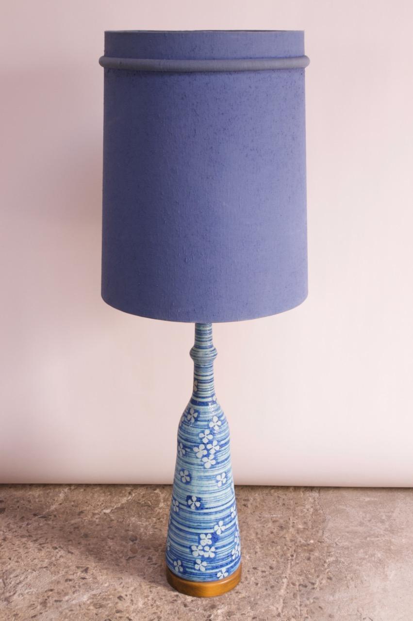 Beeindruckende Tischlampe aus Keramik in dunkel- und hellblauem Streifenmuster mit Blumenmotiv. Sie wird von einem runden Sockel aus Nussbaumholz getragen und ist mit einem großen, violetten Schirm ausgestattet. Merkmale von Raymor- und