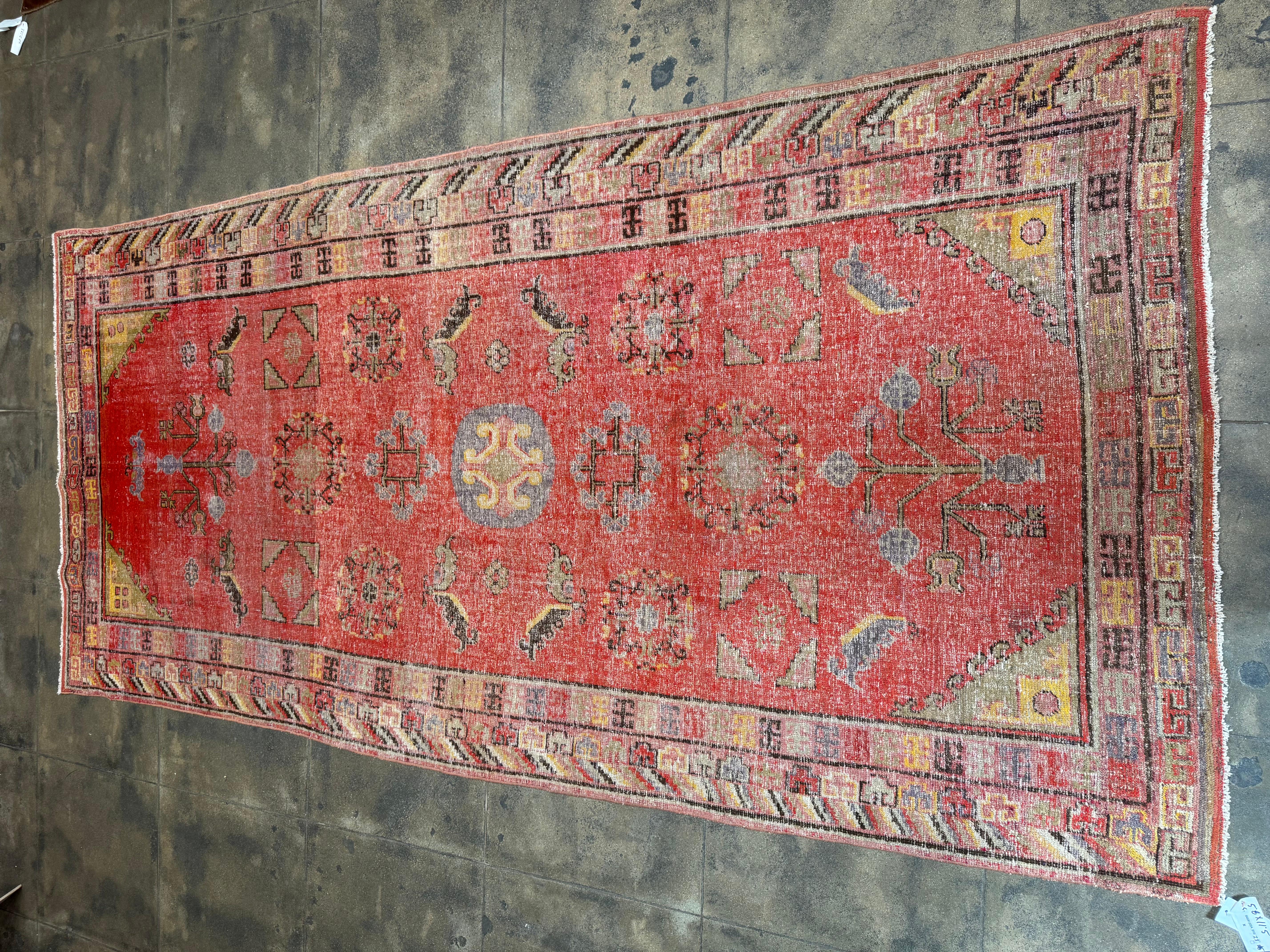 Esta alfombra Khotan Samarkand vintage de gran tamaño presenta una cautivadora estética tribal con un atrevido campo rojo, intrincados medallones geométricos y motivos simbólicos del árbol de la vida y el cuerno de carnero. Anudada a mano con el