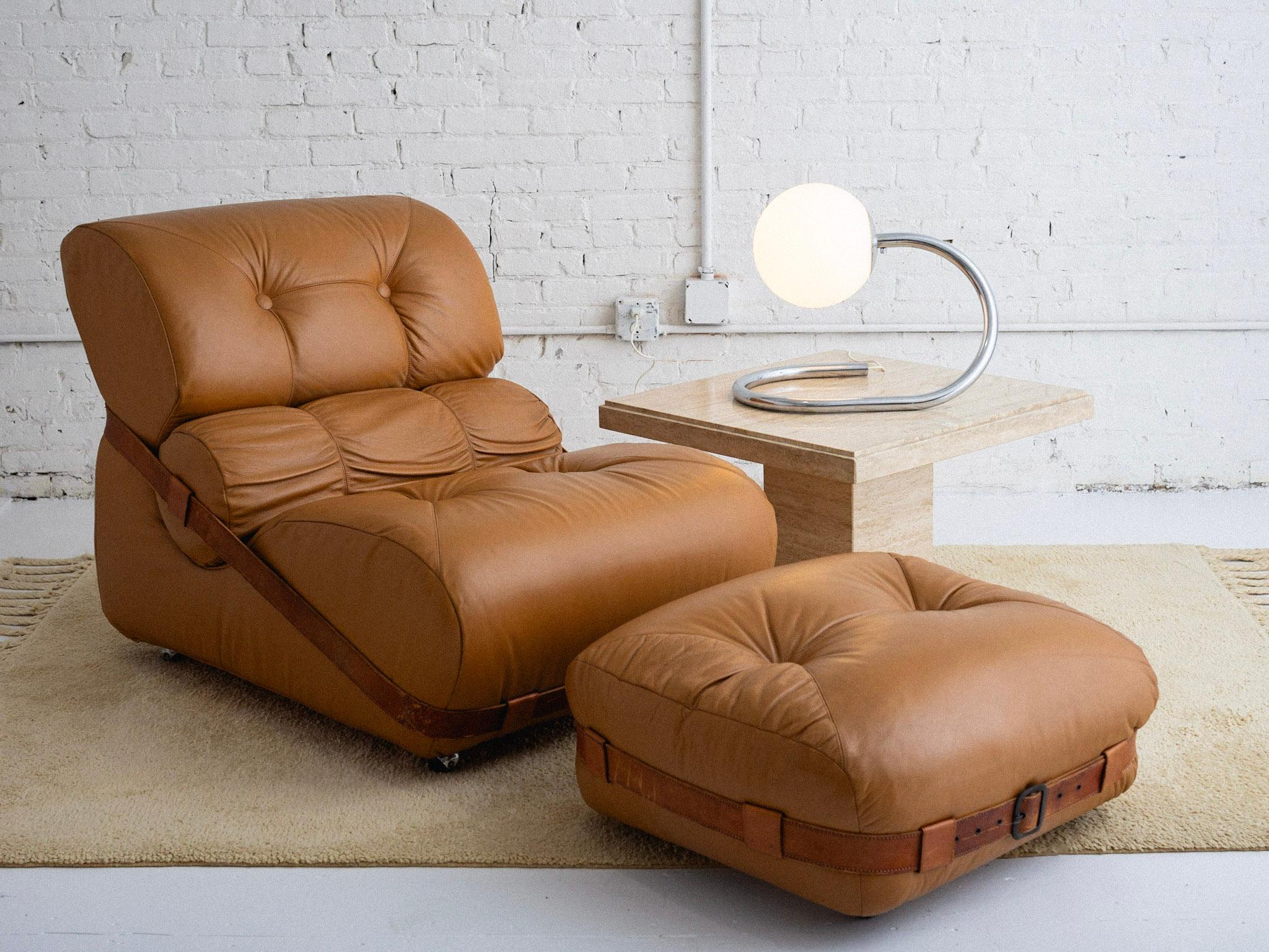 An overstuffed leather chair and ottoman in the style of De Pas, D'Urbino & Lomazzi. Camel leather chair features a back bolster with surrounding leather belt strap. Matching ottoman also features a surrounding belt. Both pieces are on castors.