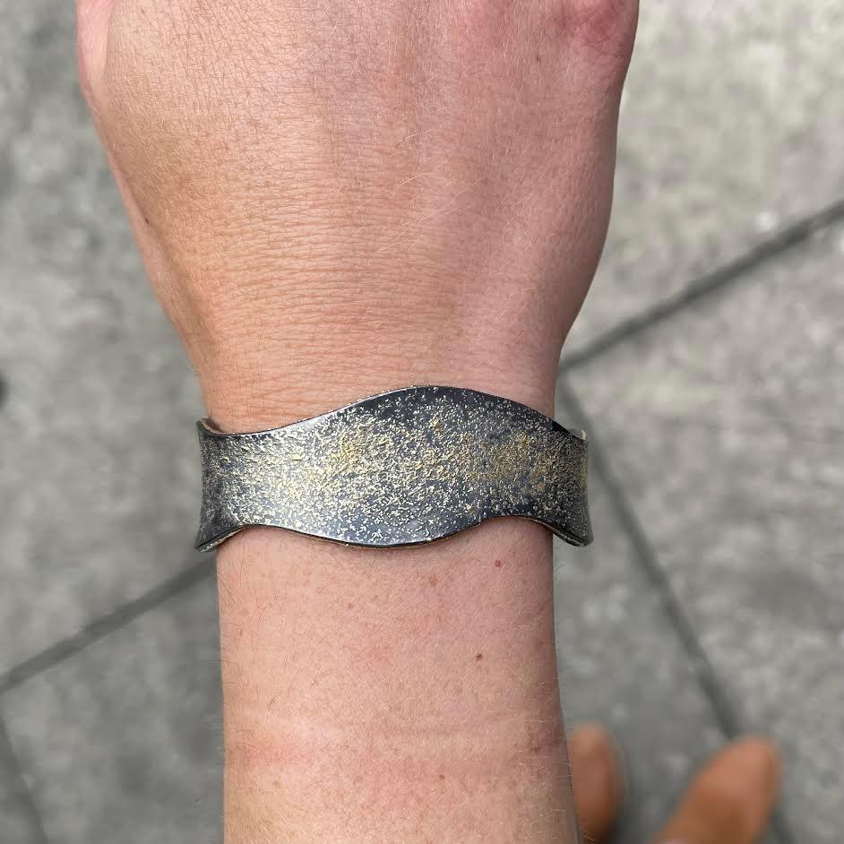 A wavy cuff bracelet made from oxidized sterling silver with 24k gold dusr fused onto the surface. This bracelet was made and designed by llyn strong.