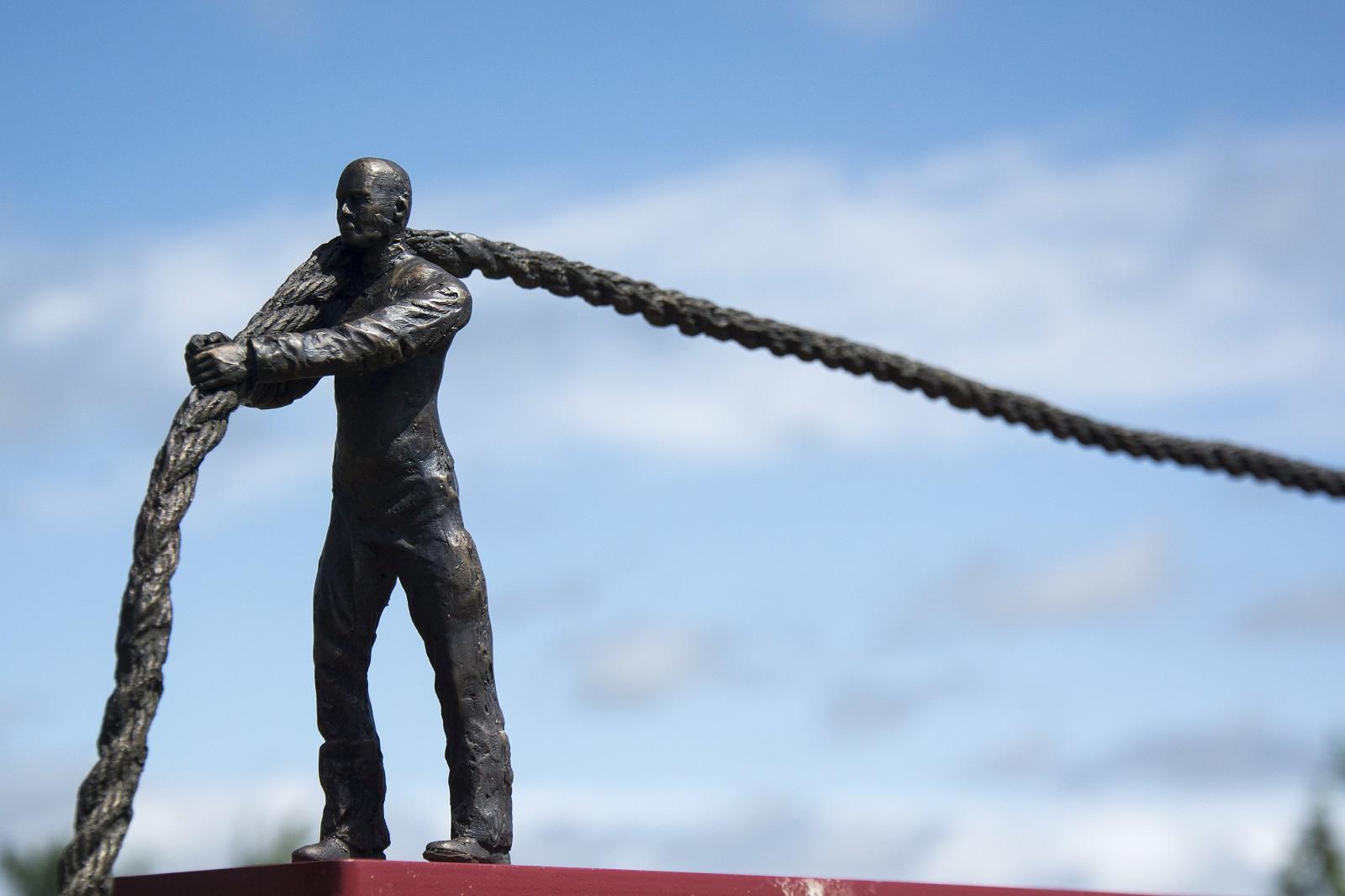 Bronze cast figurines of two men, one at each at end of a bronze rope, standing on a teeter totter  -- one man merely holds his end of the rope, the other one pulls. The whole is balanced on a black steel upright pipe. Play and playfulness are at