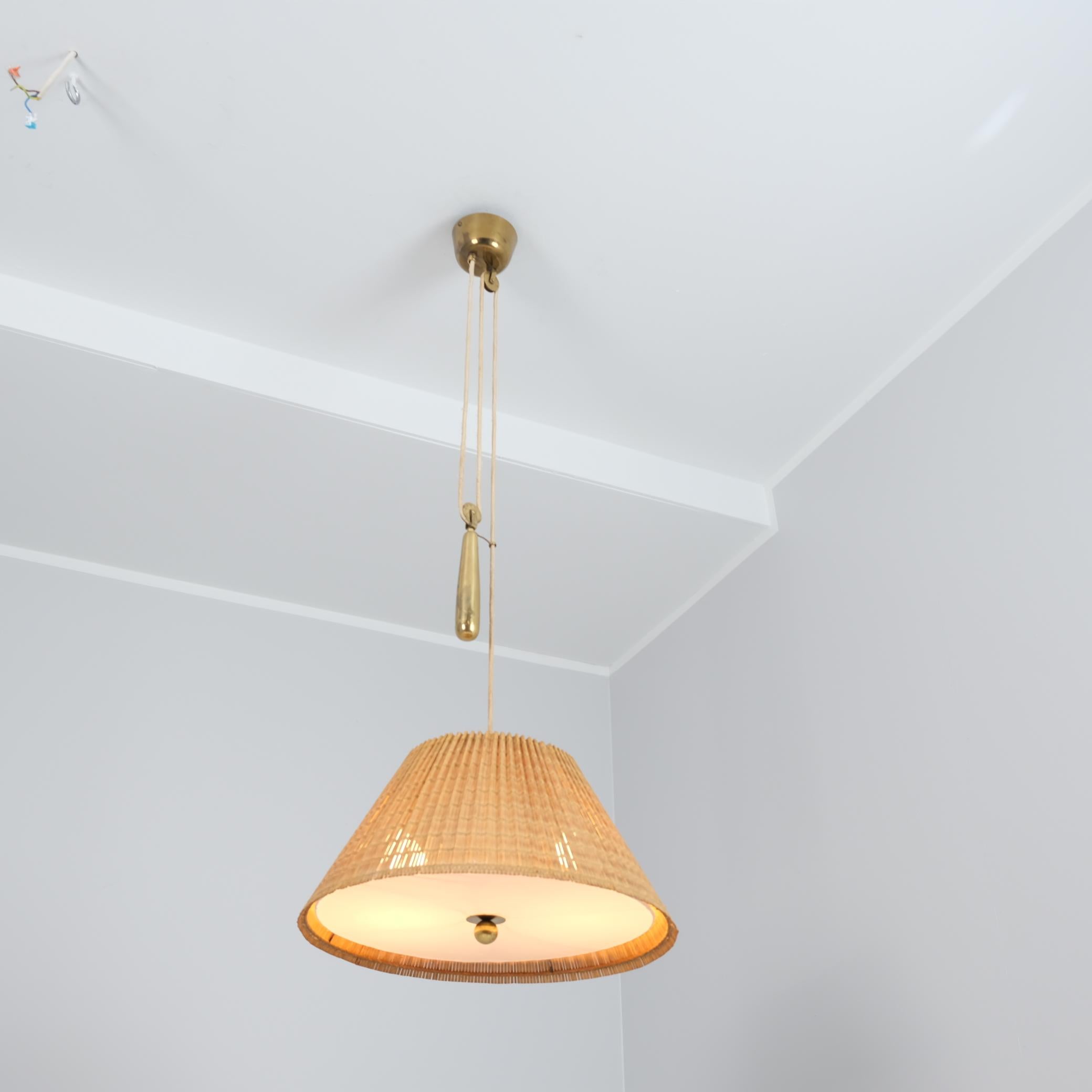 Una bellissima lampada regolabile di Paavo Tynell che unisce armoniosamente l'ottone patinato con il paralume in rattan e tessuto. Il contrappeso mette in evidenza il genio di Tynells, rendendo la lampada adatta a diverse altezze di soffitto. La