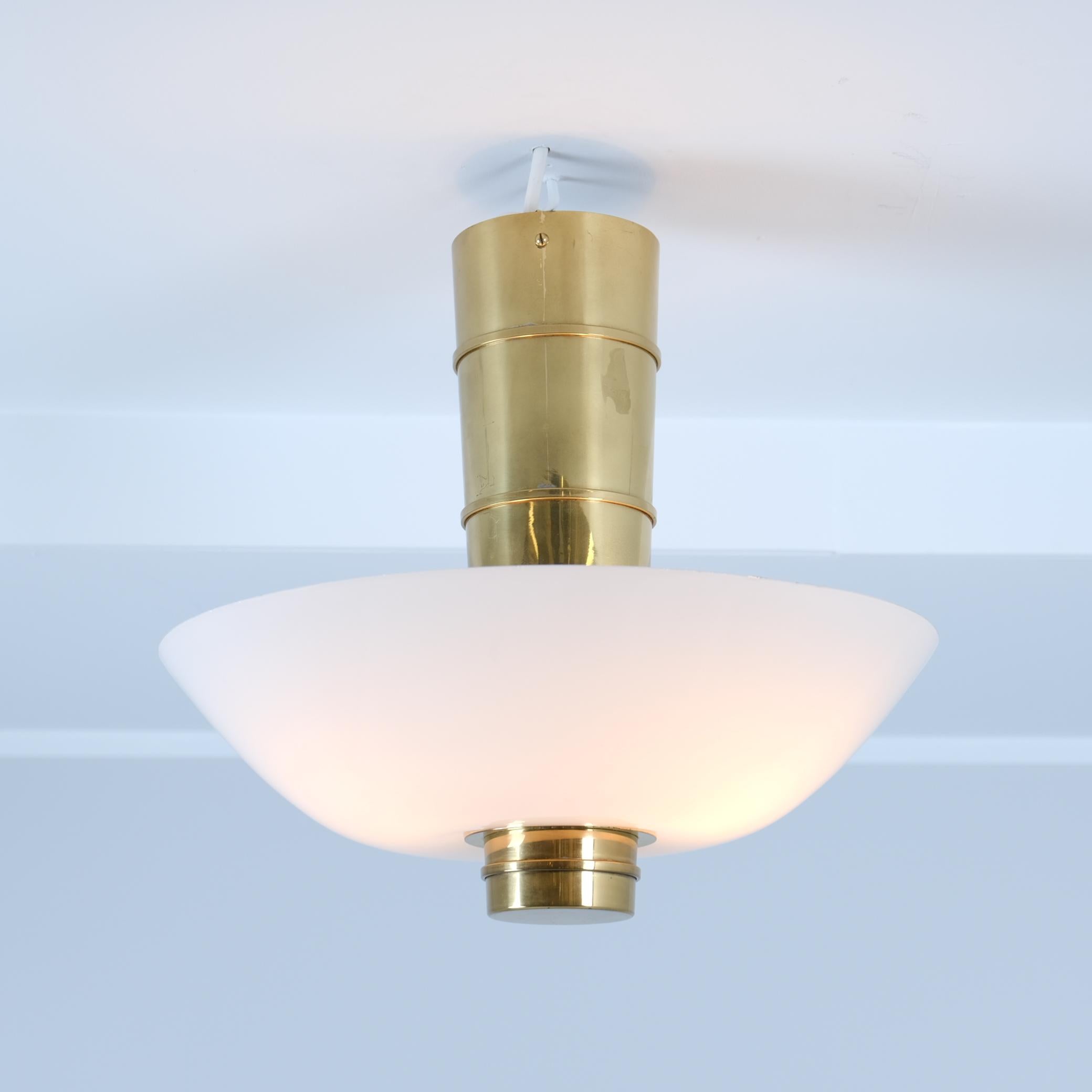 Un magnifique plafonnier de Paavo Tynell, surnommé l'obus de canon en raison de sa grande ressemblance. Élégante et minimaliste, elle associe un centre en laiton à un abat-jour en verre opalin permettant à la lumière de passer à travers mais aussi
