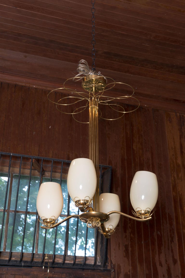 A whimsical and elegant brass and glass chandelier in the style Paavo Tynell. Four oval blown glass shades. Brass loop wire decoration on the canopy. Large loops just below the canopy. Loops on the brass bobeches. Circular loops surrounding a ball