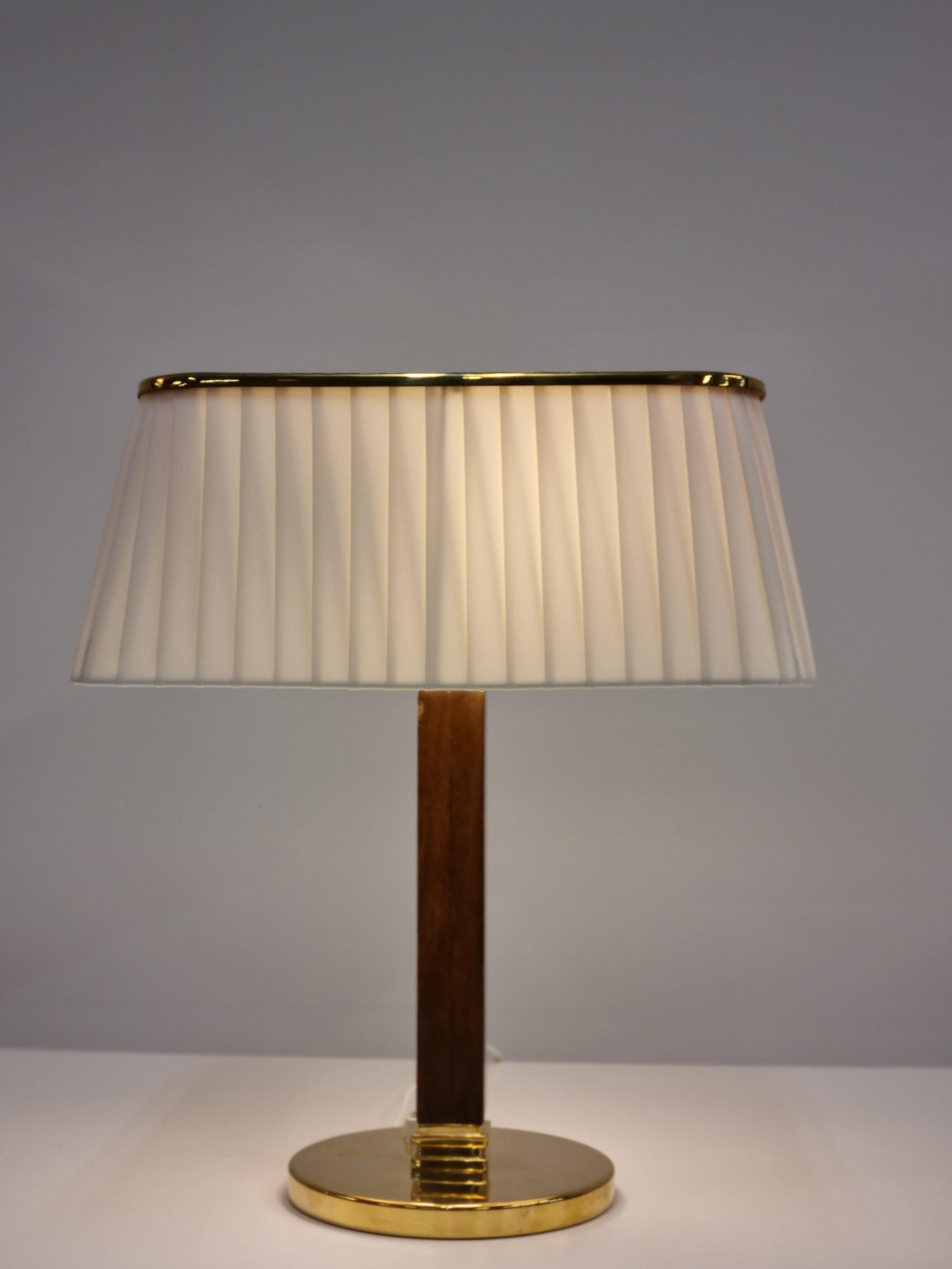 This table lamp model 5066 by Paavo Tynell for Taito Oy from 1950s is a classic. It has a brass round stand, a hard wooden leg and a beautiful light colored pleated textile shade. The square brass shade top has a fibonacci sequence perforation and