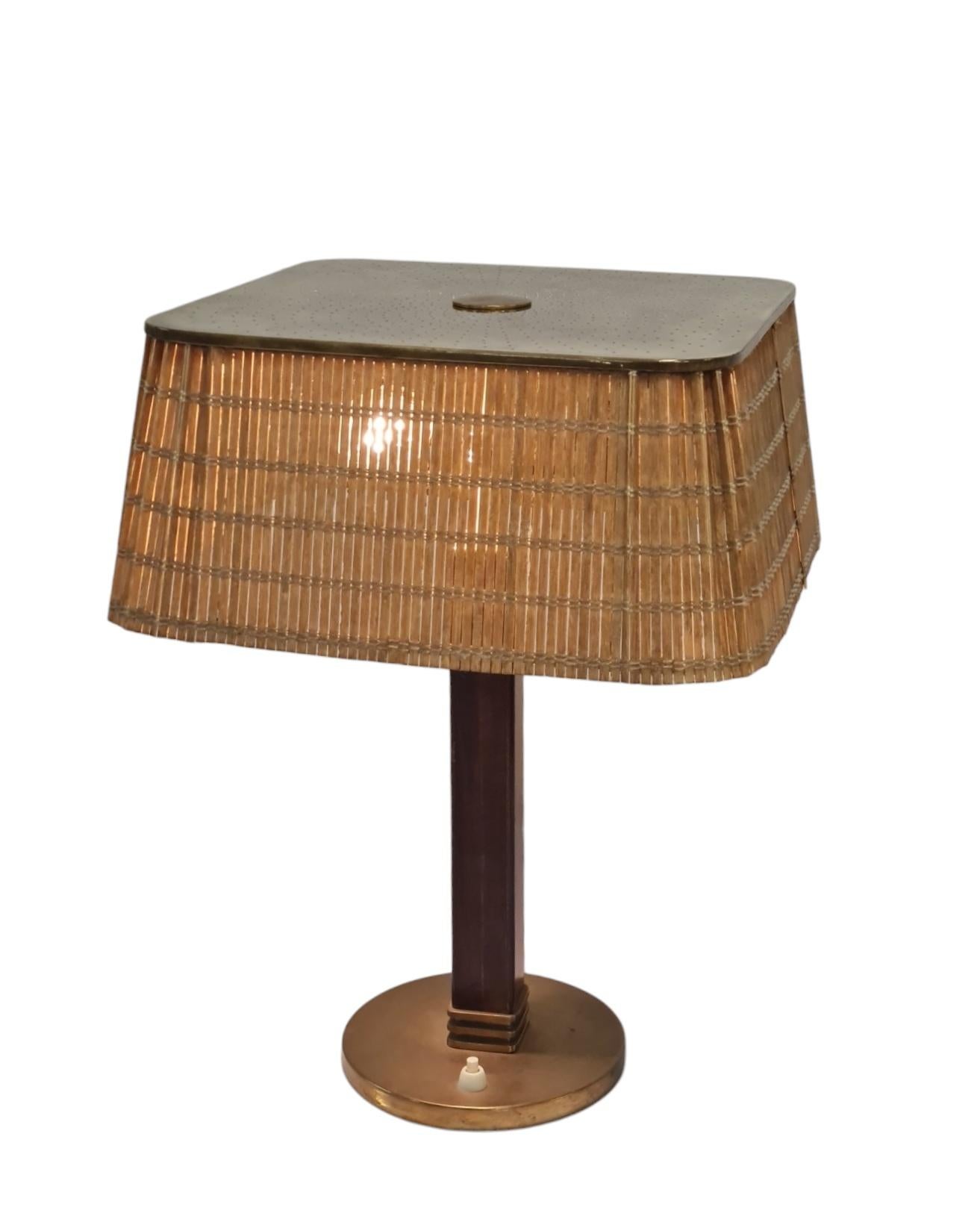 This table lamp with model no.5066 by Paavo Tynell for Taito Oy from the 1950s is a classic. 
It has a brass round stand, a hard wooden leg and a beautiful clean lined light mahogany color rattan shade. The square brass shade top has a fibonacci