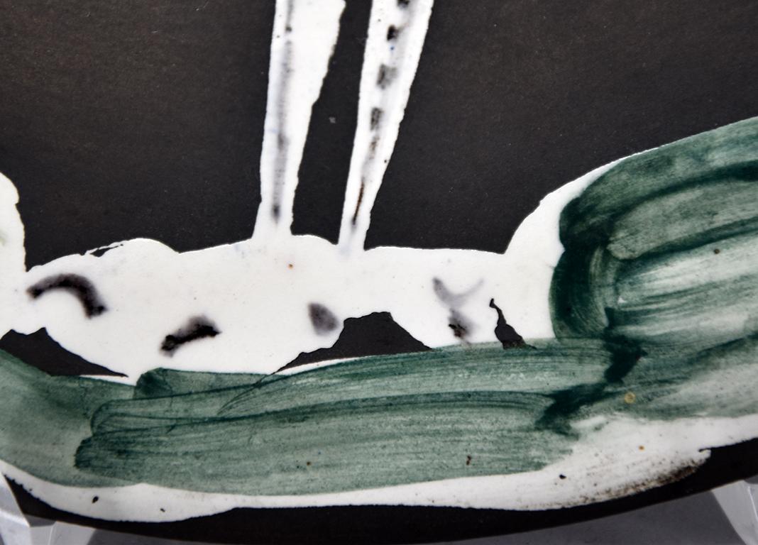 Créée en 1963, cette assiette en terre cuite madoura à décor en engobe (vert, blanc, noir) et émail sous glaçure partielle brossée, patine noire est inscrite sur le fond 