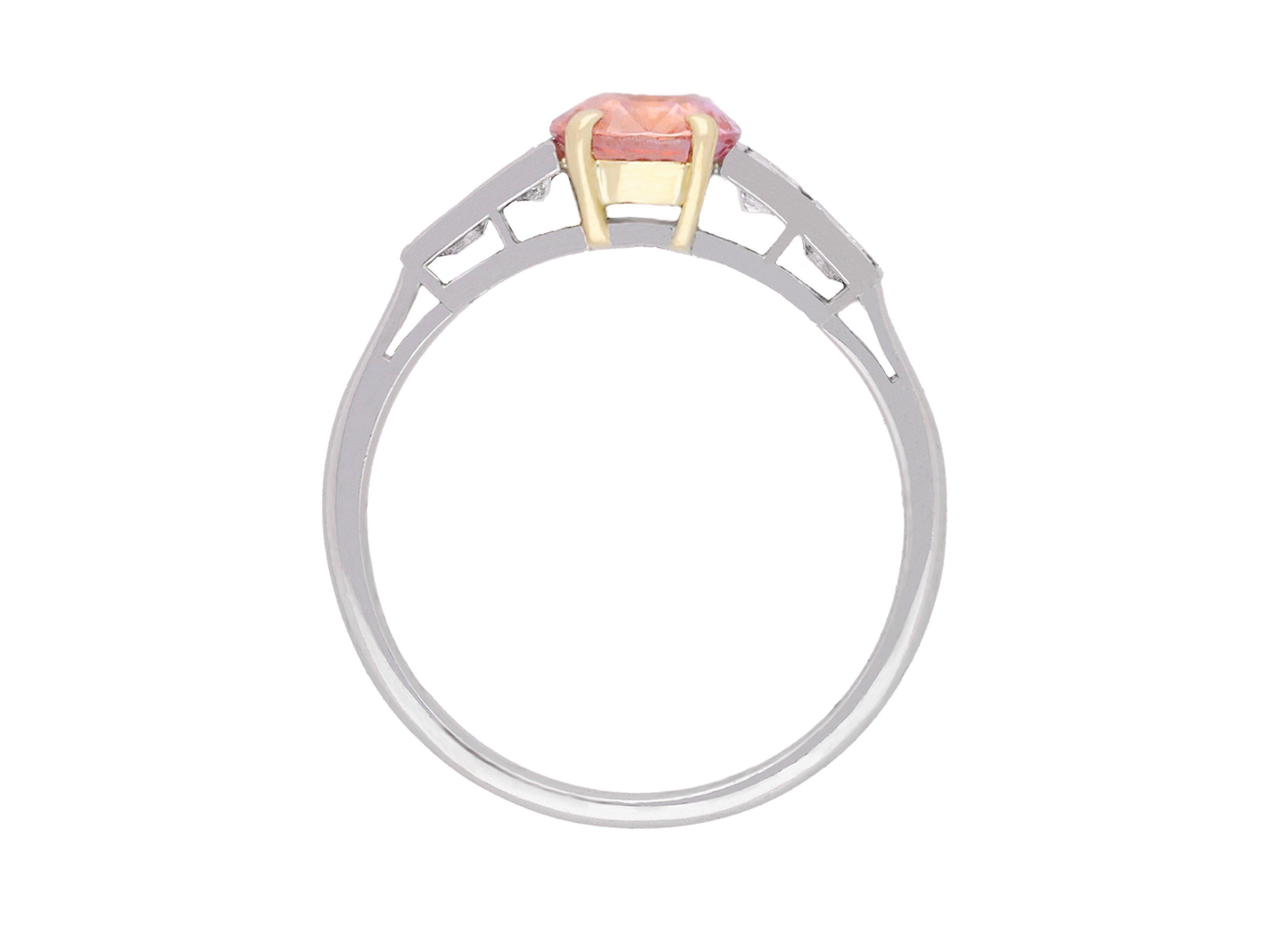 Padparadscha Ceylon sapphire and diamond ring. Set to centre with a cushion shape mixed cut natural unenhanced Padparadscha Ceylon sapphire in an open back claw setting with a weight of 1.18 carats, flanked by two tapering baguette cut diamonds in