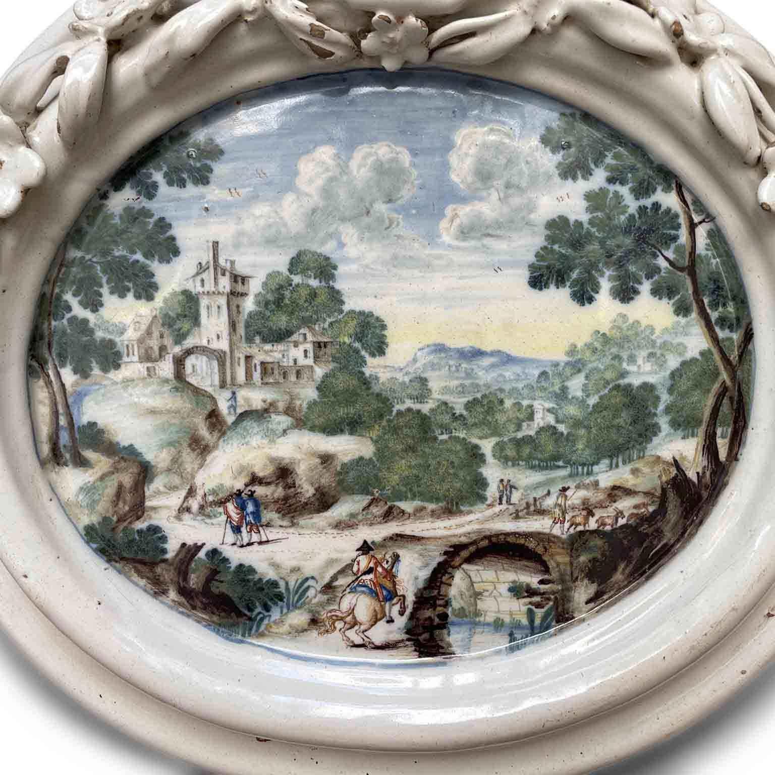 Italienische polychrome Maiolica-Wandfliese, venezianischer Bereich mit Landschaft bemalt 1750 Circa,  oval mit einem Rahmen, einem aus Blüten und Trauben gebildeten Cymatium, das von einer Muschel überragt wird.  Diese geformte Majolika-Plakette