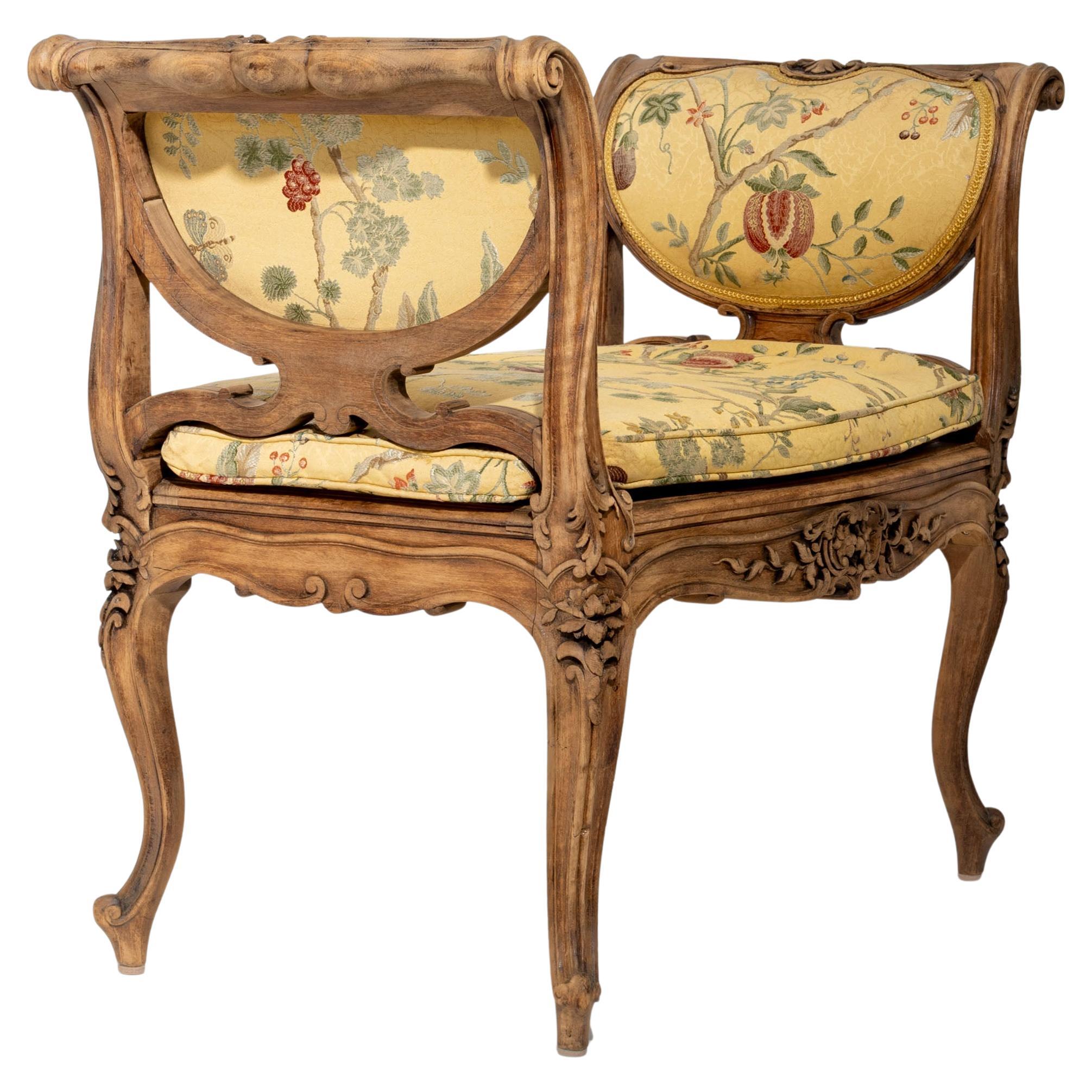 Small wooden page bench or stool with serpentine apron and floral carved decoration. The voluted side armrests curl elegantly outward. Upholstered in a cheerful yellow fabric featuring branch and pomegranate motifs. A stylish 20th-century