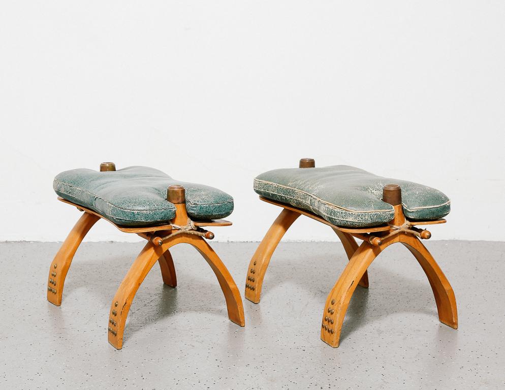 Camel saddle stools with teal vinyl upholstery. Legs joined to seat with leather string. Brass accents.