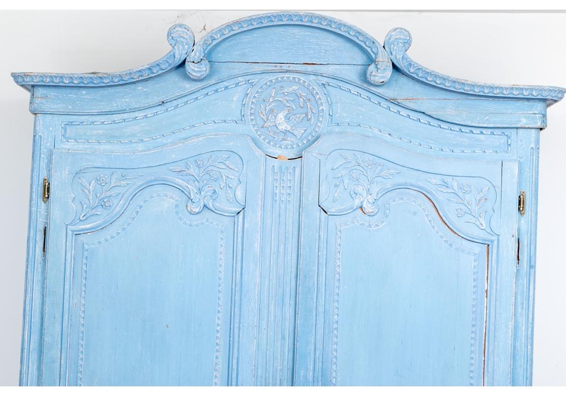 Paint decorated 19th century French cornflower blue armoire in three parts. The armoire with two doors having brass escutcheon plates, recessed cartouche form panels separated by musical heraldic devices within egg/dart circles. The armoire with a