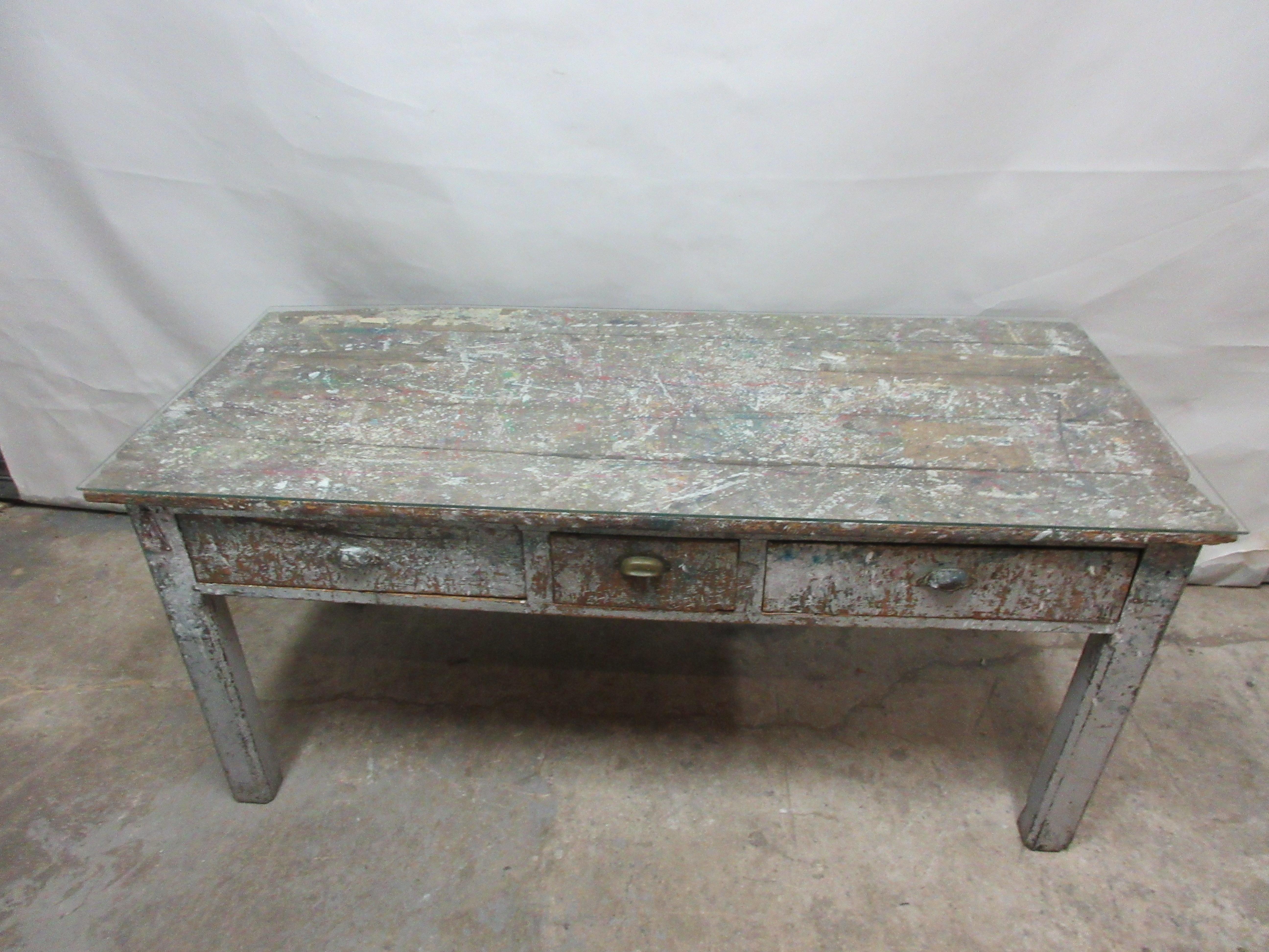 This is an antique work shop paint table with 3 drawers. It has a glass top.