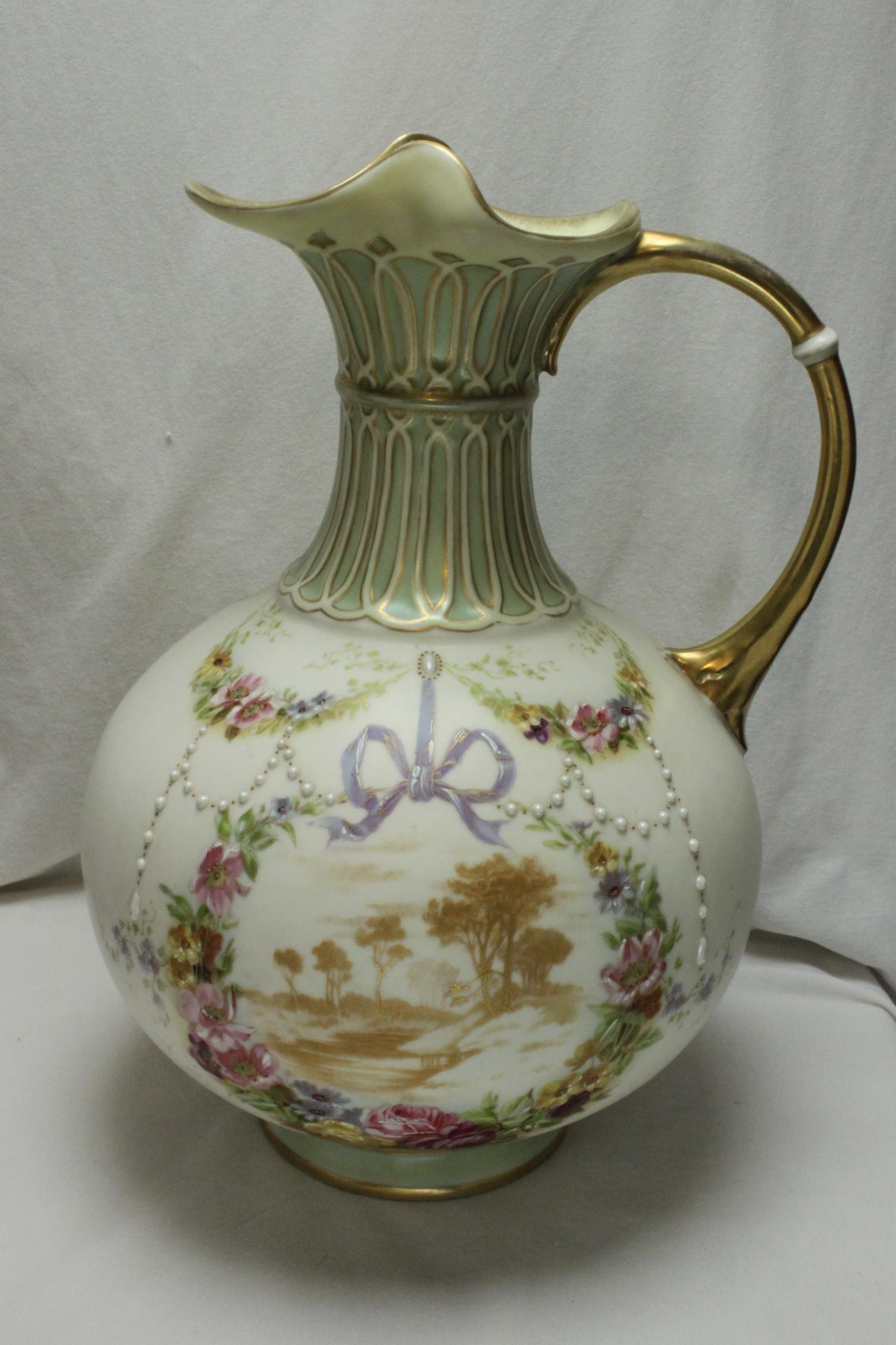 This very pretty painted and gilded parian jug or ewer, is decorated with hand painted ribbons, swags of jewelled and highlighted flowers and strings of jewelled pearls. In the centre is a rural scene, printed in gold and encircled by a cartouche of