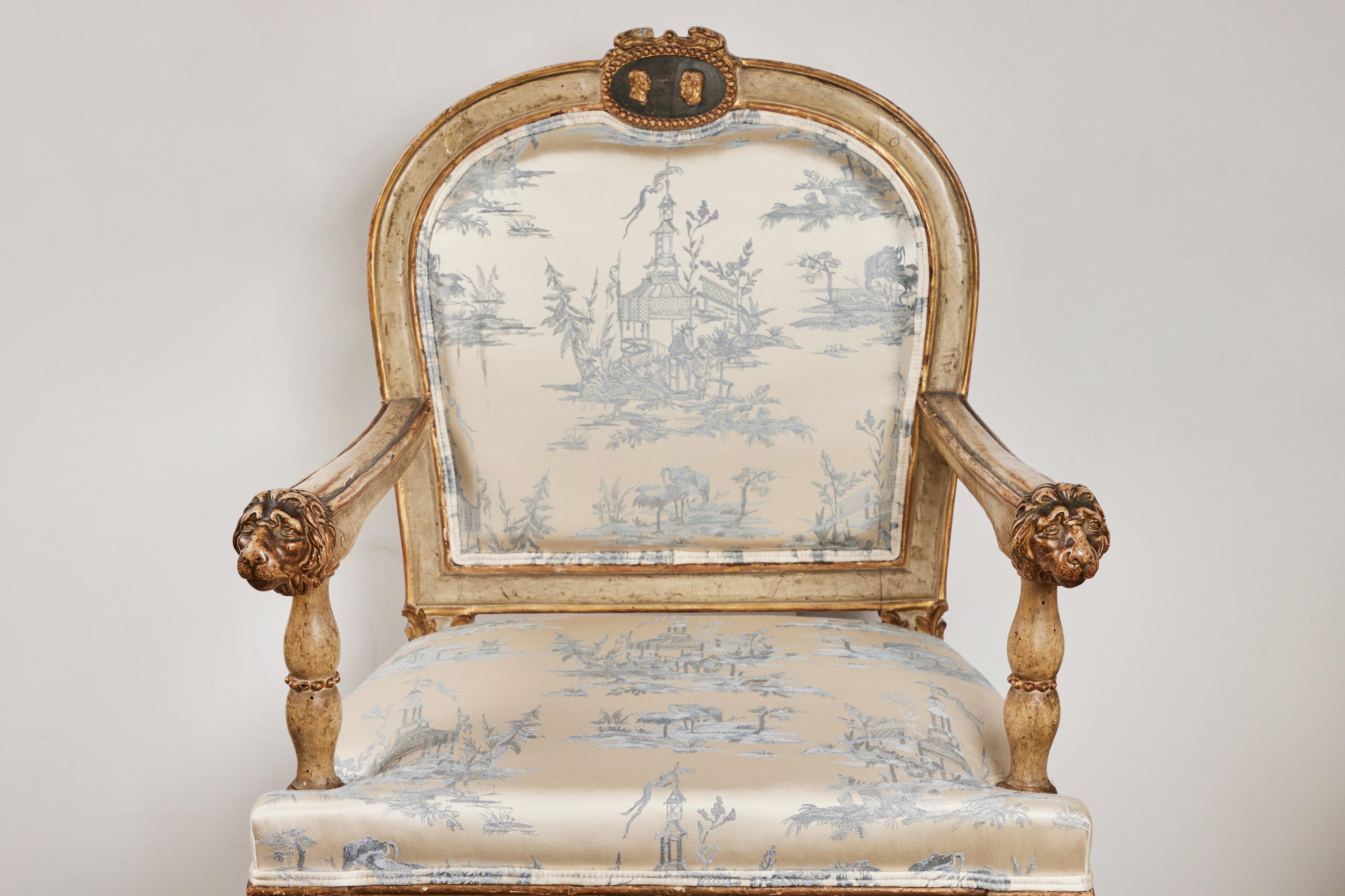 Hand carved, painted and parcel gilt armchair from the area of Rome. Design features include lions head on arms, facing male profiles and bird reliefs, beaded pattern and fluted legs. New Silk Chinoiserie themed upholstery.