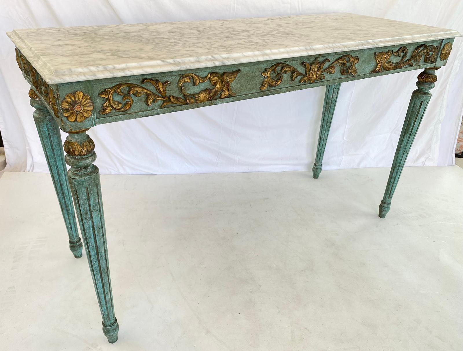 Console table, in the Louis XVI style, having a rectangular top of Carrara marble, its base painted in teal, showing natural wear, its apron decorated with parcel gilt foliate scrollwork, flanked by rosette blocks, raised on round, tapering, fluted