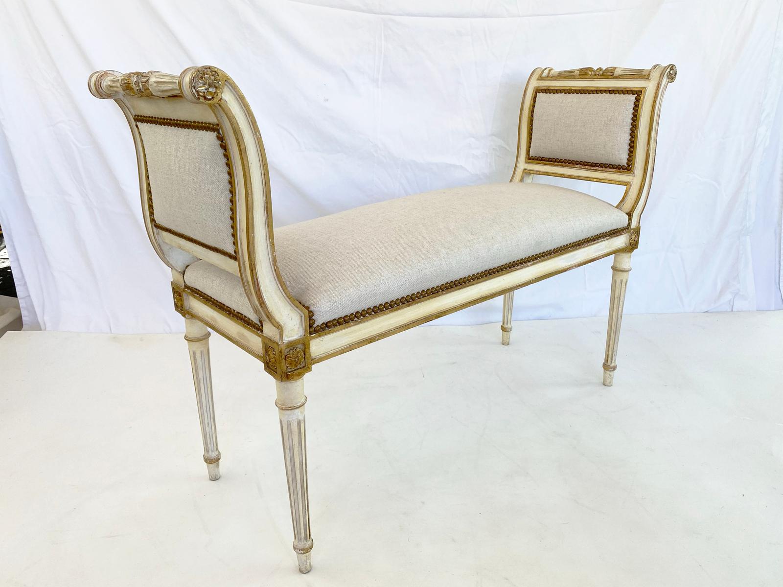 Window seat bench, painted and parcel gilt finish showing natural wear, having a fielded frame, with padded outscrolled arms finished with a rosette, and turned and reeded handle, padded seat, in linen, finished with nailheads, raised on round,