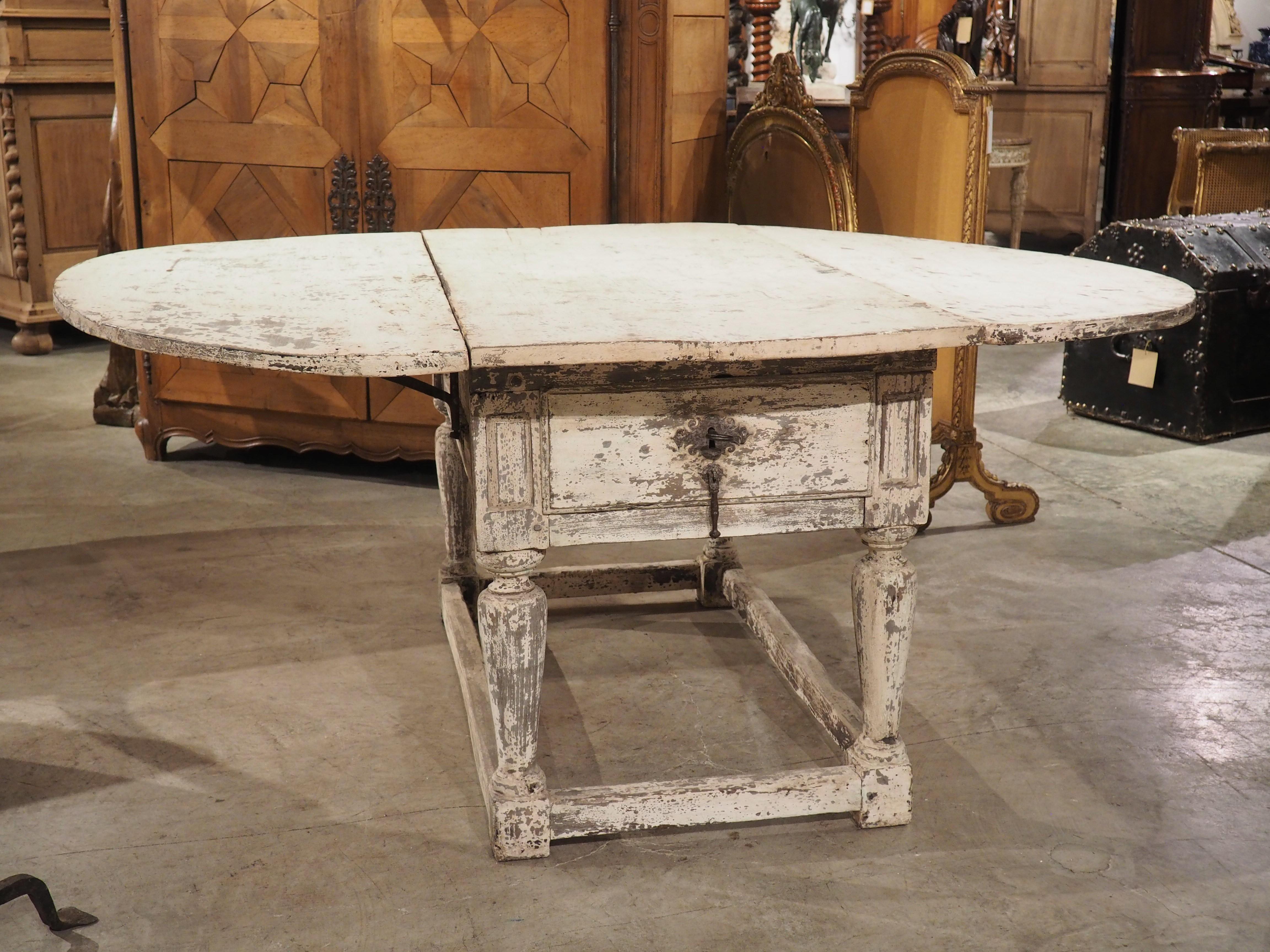 A wonderful and unique table from Italy, this painted oval drop-leaf table has two drawers on the long sides, both with functioning locks (unlocked by a single key).  The oak was hand-carved in the 1600s, with the distressed white finish being added