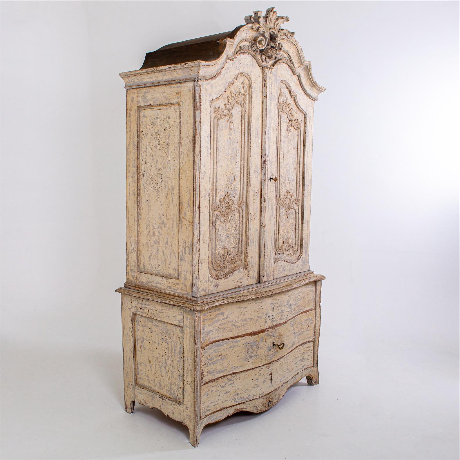 Large baroque cabinet with three-drawer commode base on a curved frame and two-door cabinet with crowning carved volute decoration and curved fillings. The setting is beige and was subsequently patinated with an antique finish.