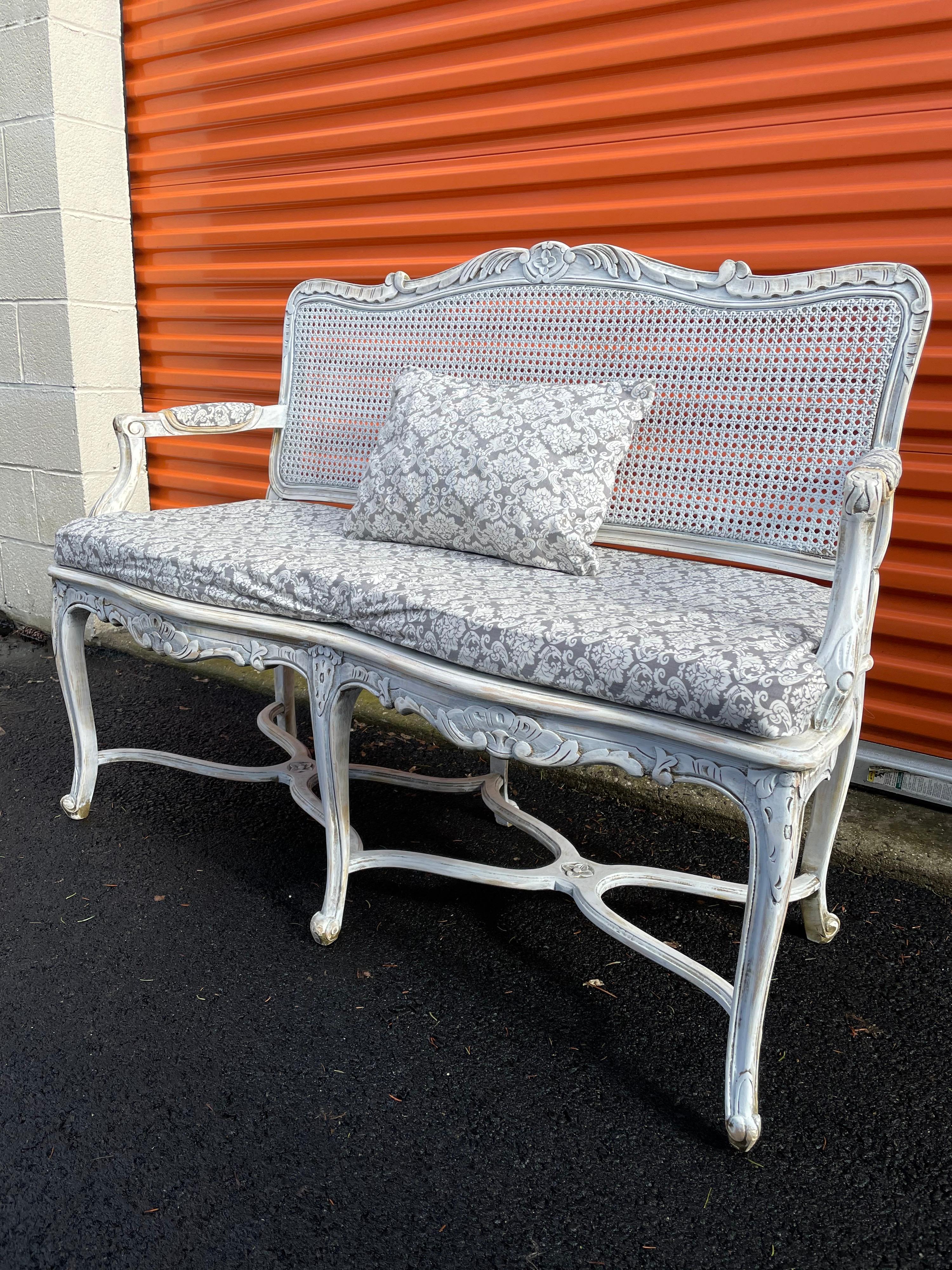 Painted carved caned bench. Shown with upholstery wrapped around original silk cushion on caned bottom. Arms have matching upholstery, comes with small matching pillow.